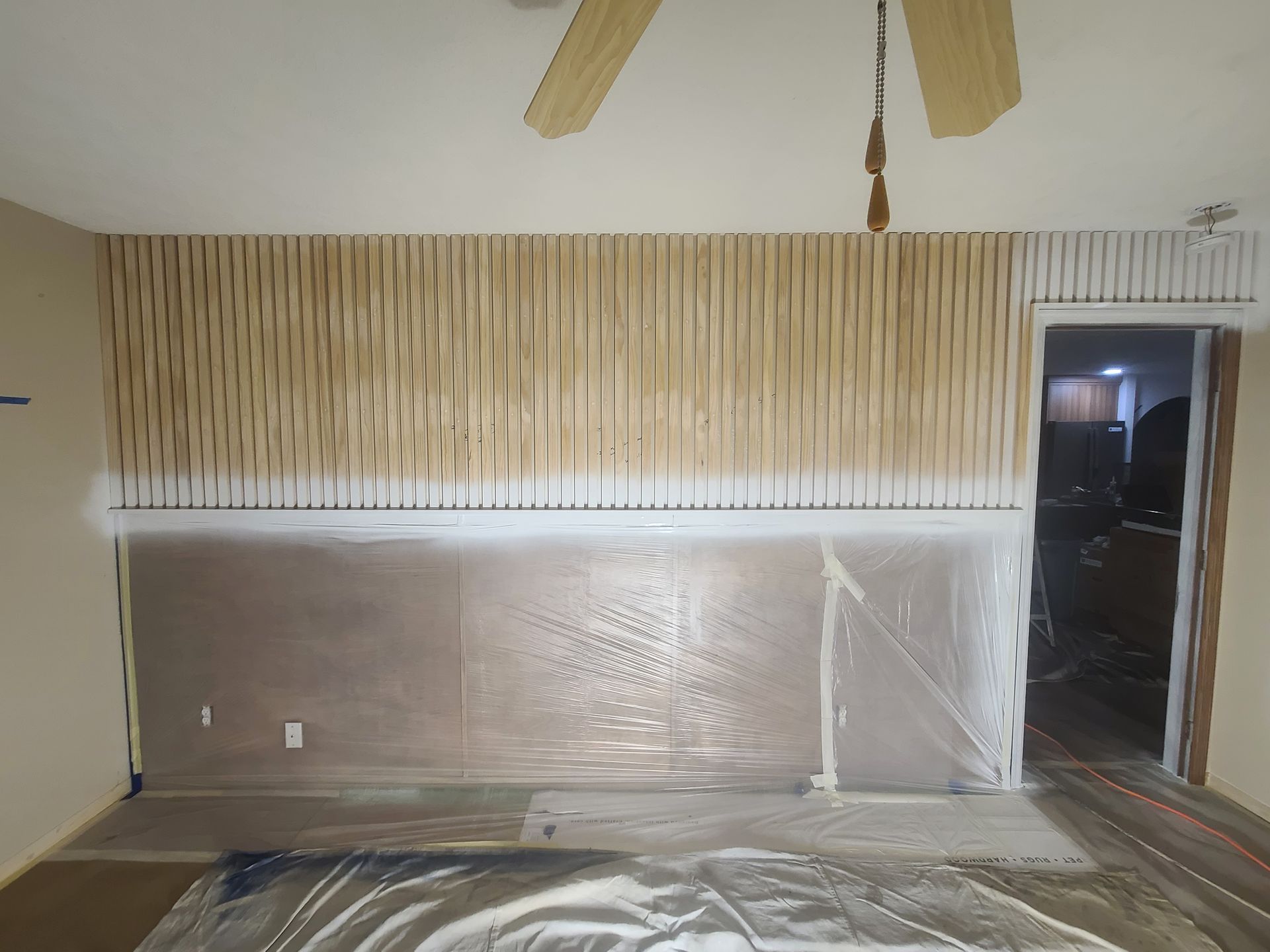 Room with wood slat wall, white trim, plastic sheeting on floor, and open doorway.