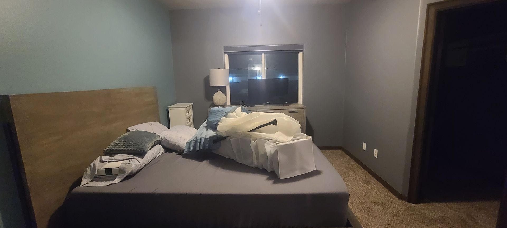 A cluttered bedroom with a bed, nightstand, window, and door. Gray walls, brown door frame, and carpet.