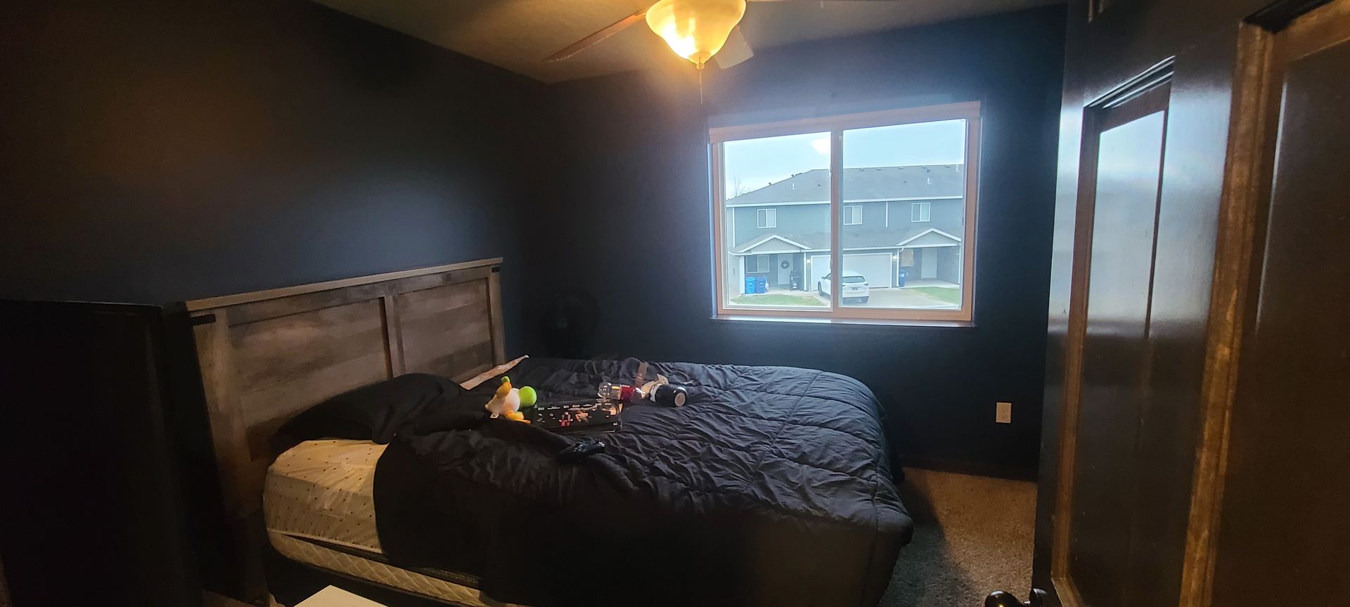 Bedroom with a bed, a window with houses visible outside, and a ceiling fan. Walls are painted dark.