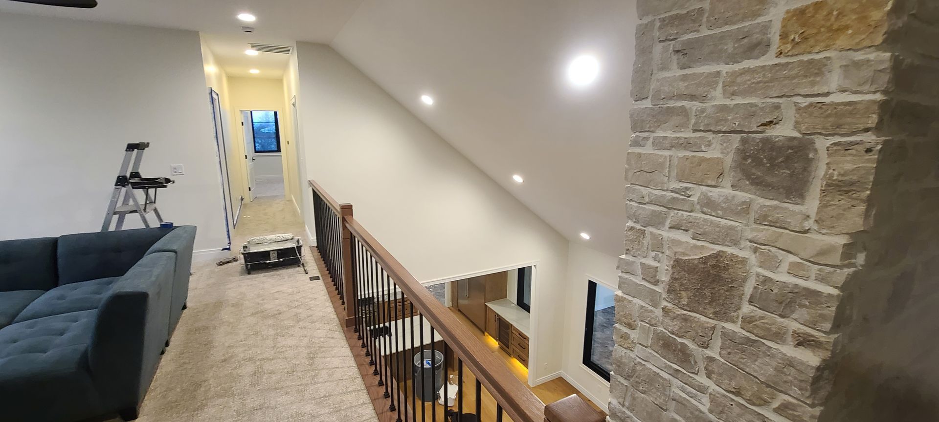 Interior view of a hallway with a stone wall and a wooden railing. There is a couch and a ladder in the view.