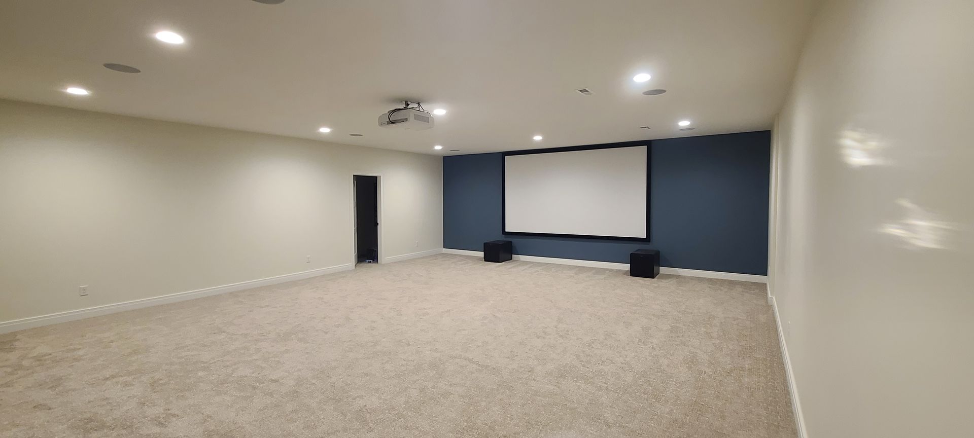 Empty home theater room with projector, screen, and blue accent wall. Carpeted floor, recessed lighting.