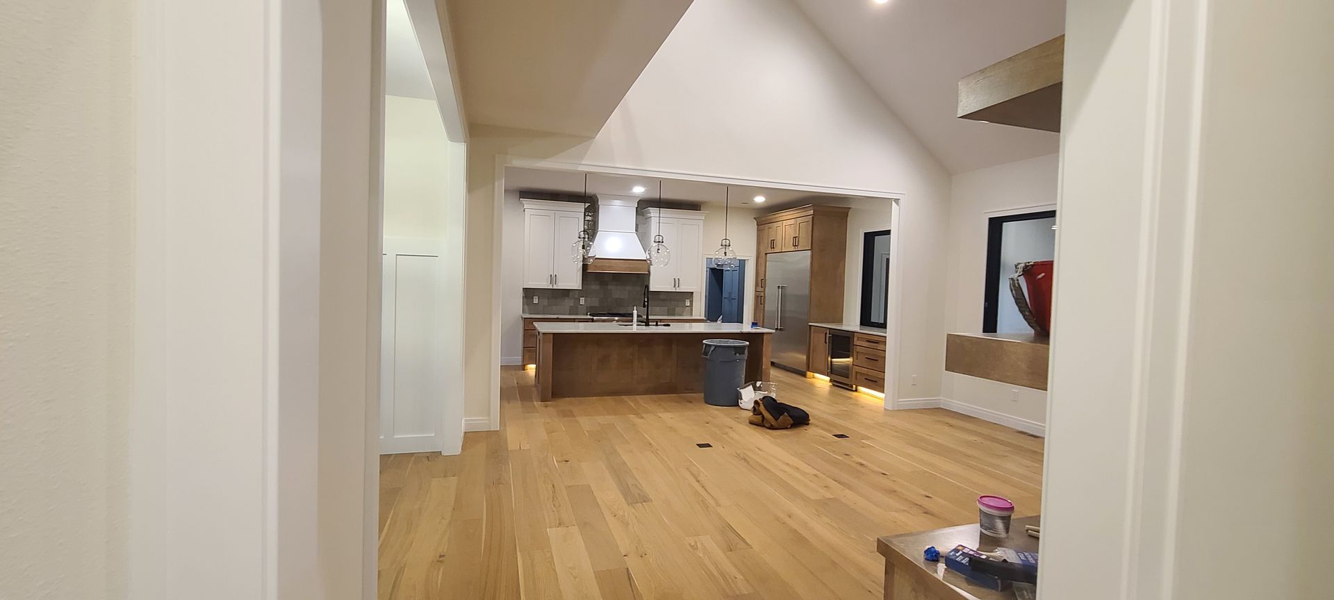 Interior view of a modern house, featuring a kitchen and living area with wood flooring and high ceilings.
