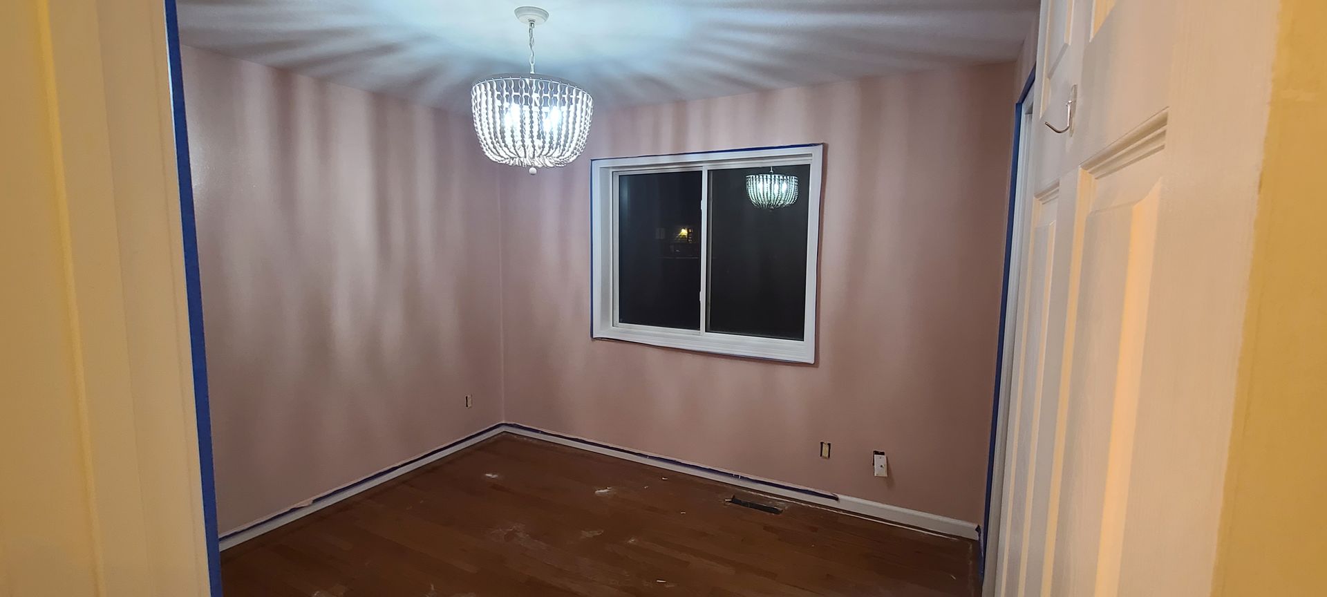 A room with pink walls, hardwood floors, and a crystal chandelier. There is a window and a door.