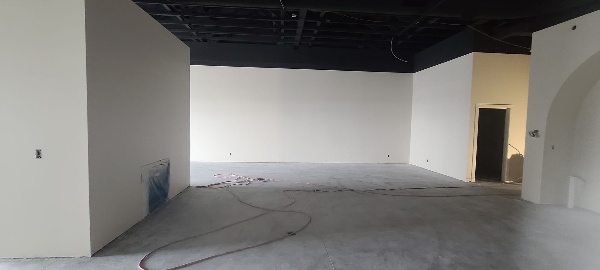 An empty room with white walls and a black ceiling. A door and an arch are visible.