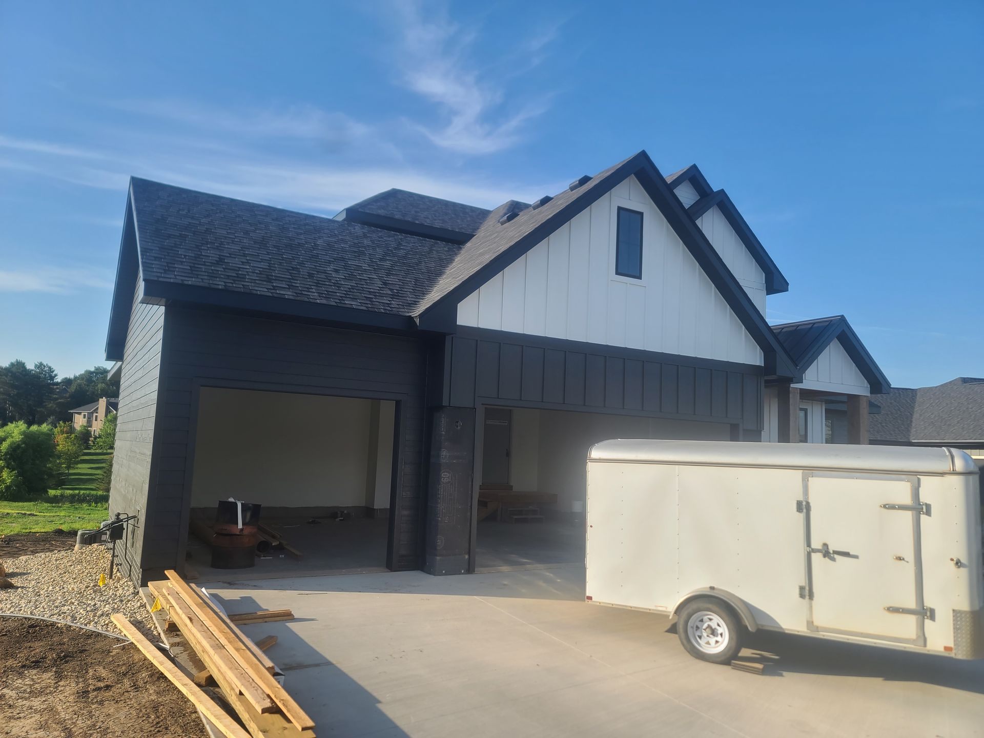 Modern house under construction with black and white exterior, open garage, trailer, and blue sky.