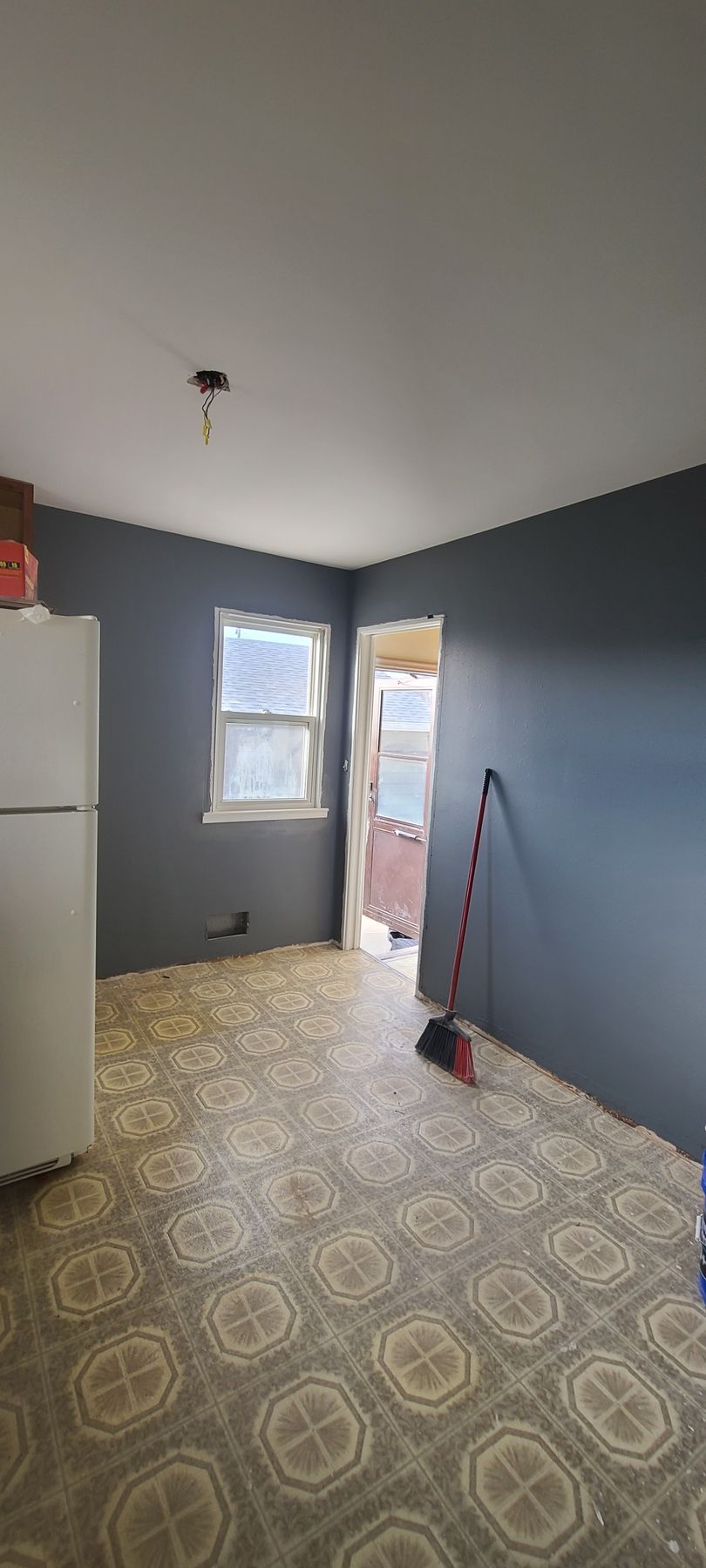 An empty kitchen with gray walls, beige patterned floor, and a white refrigerator. A door leads outside.