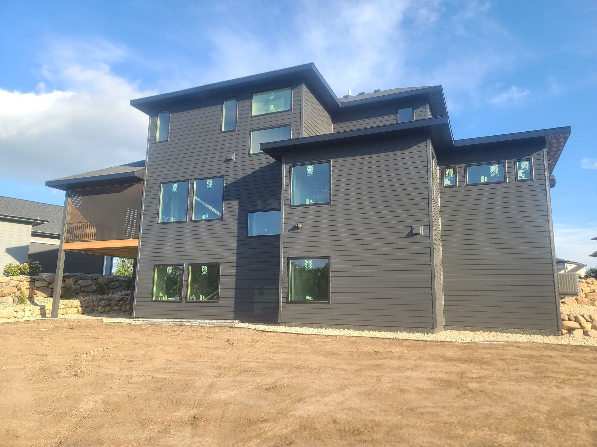 Modern two-story house with dark gray siding, multiple windows, and a flat roof against a blue sky.