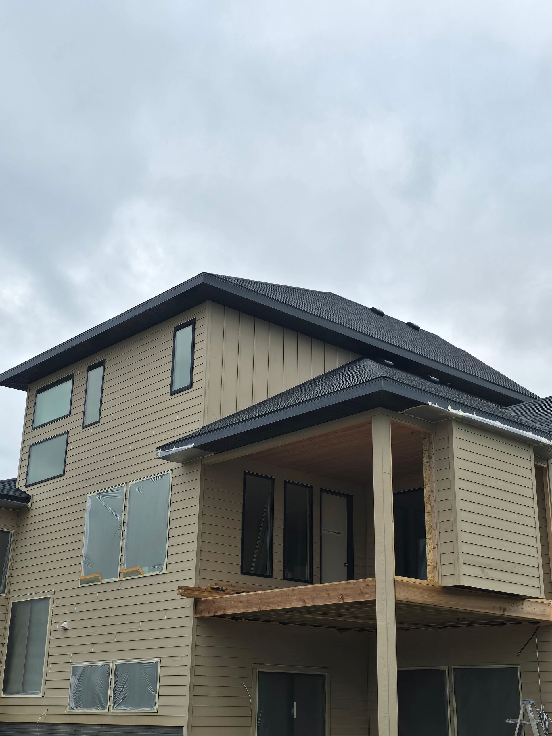 Two-story house with tan siding, black roof and trim, under a cloudy sky.