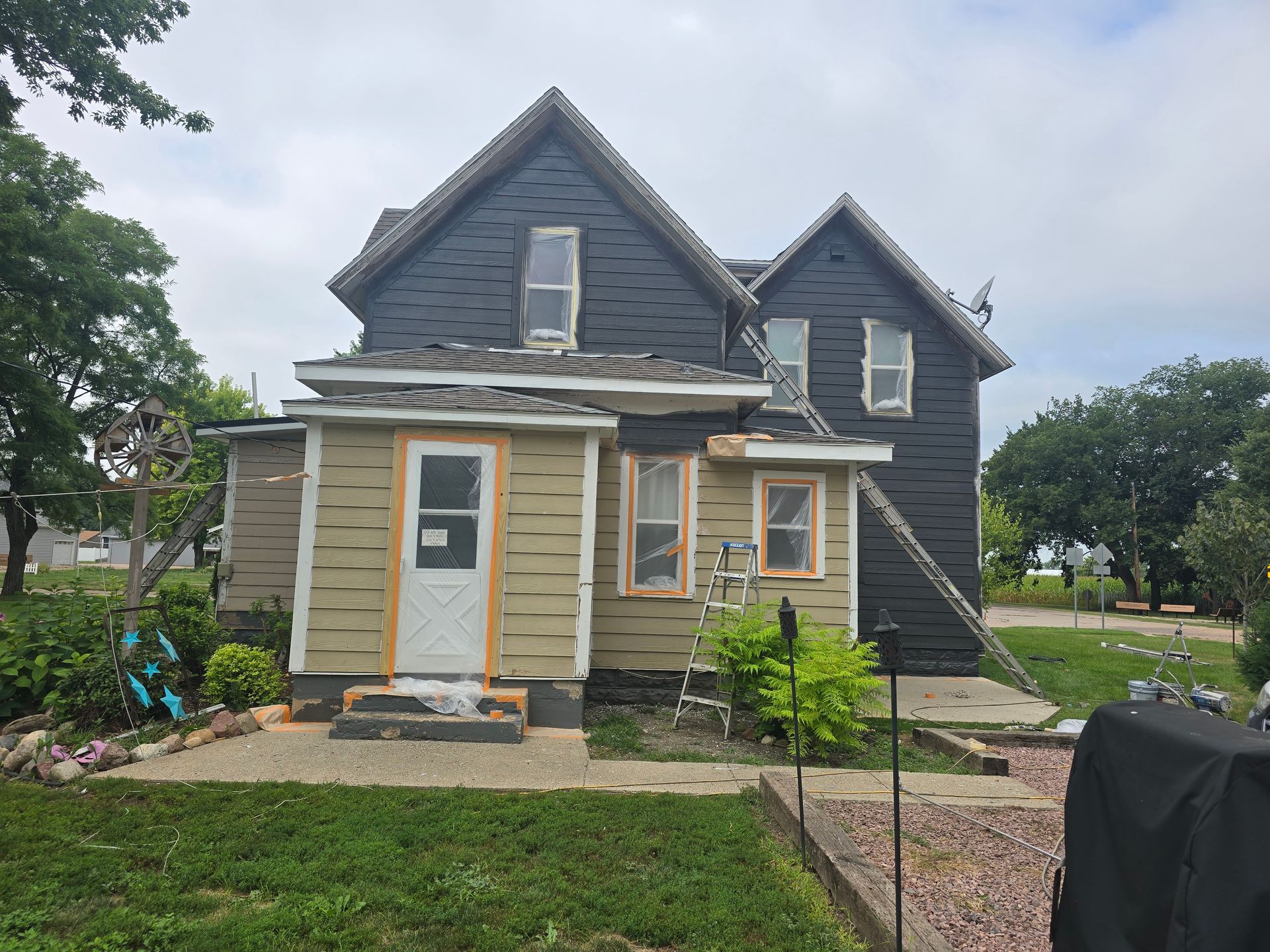 House exterior being renovated; dark gray siding, light green trim, ladders, overcast sky.
