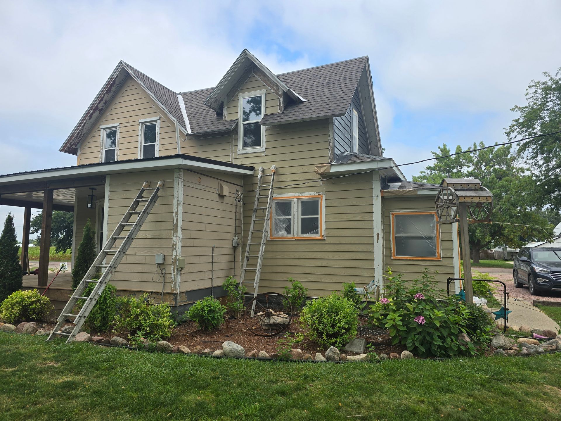 Two-story house with tan siding, porch, and ladders. Bushes and lawn in the foreground; sky in the background.