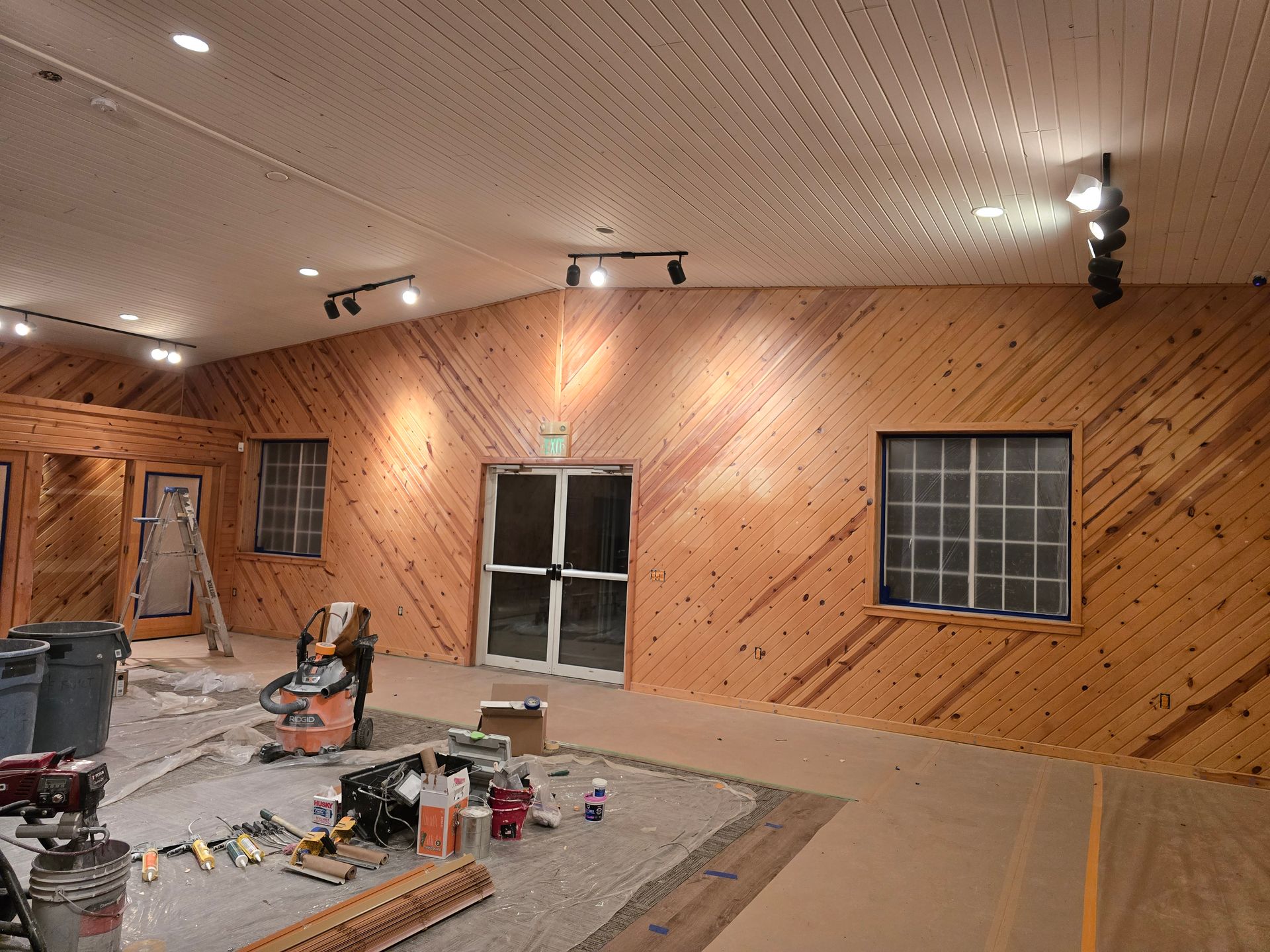 Interior room with wooden paneled walls, ceiling lights, windows, and construction materials on the floor.
