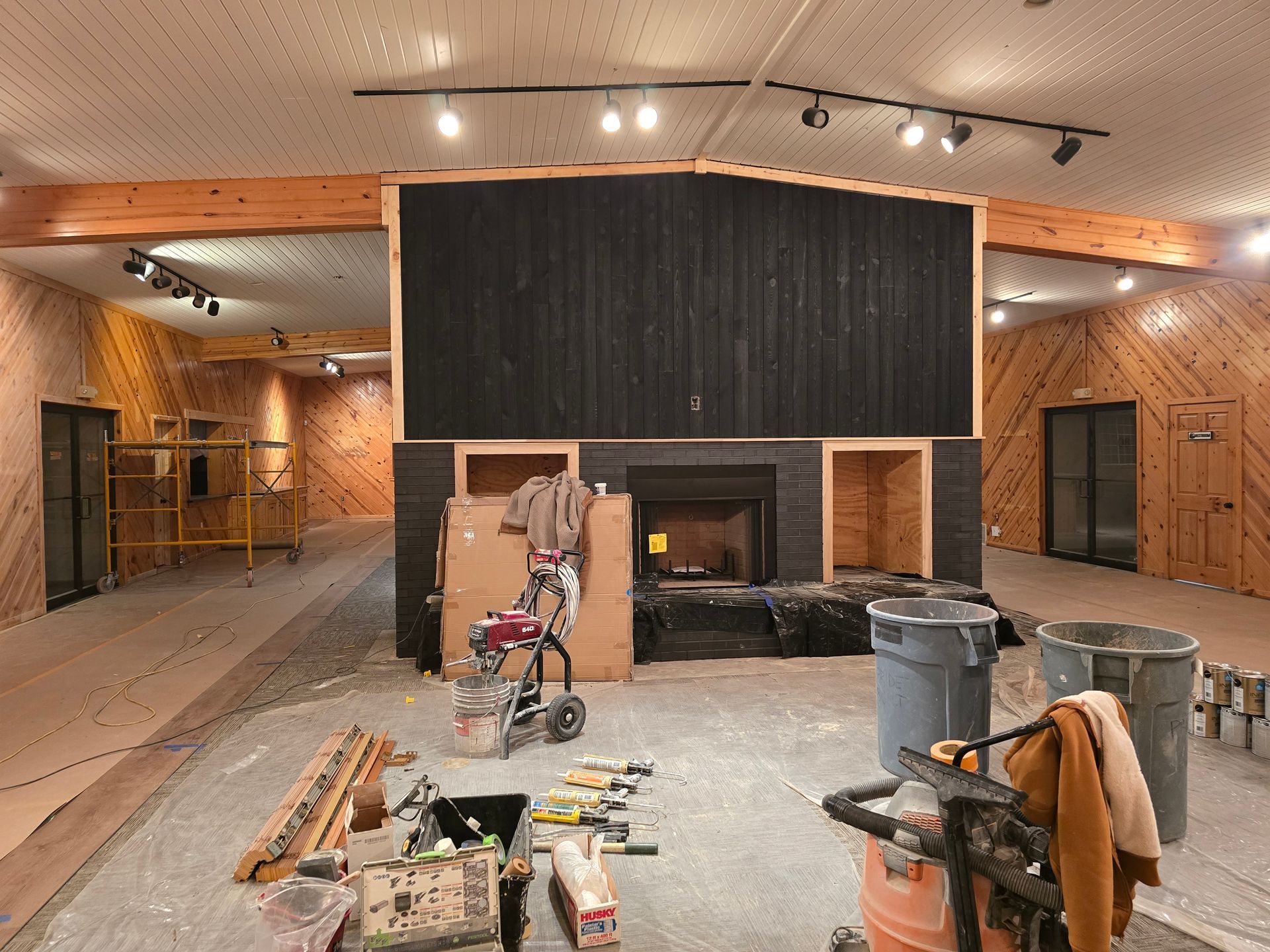 Interior renovation in progress. Fireplace with black surround, tools, wood paneling, exposed beams, and a concrete floor.