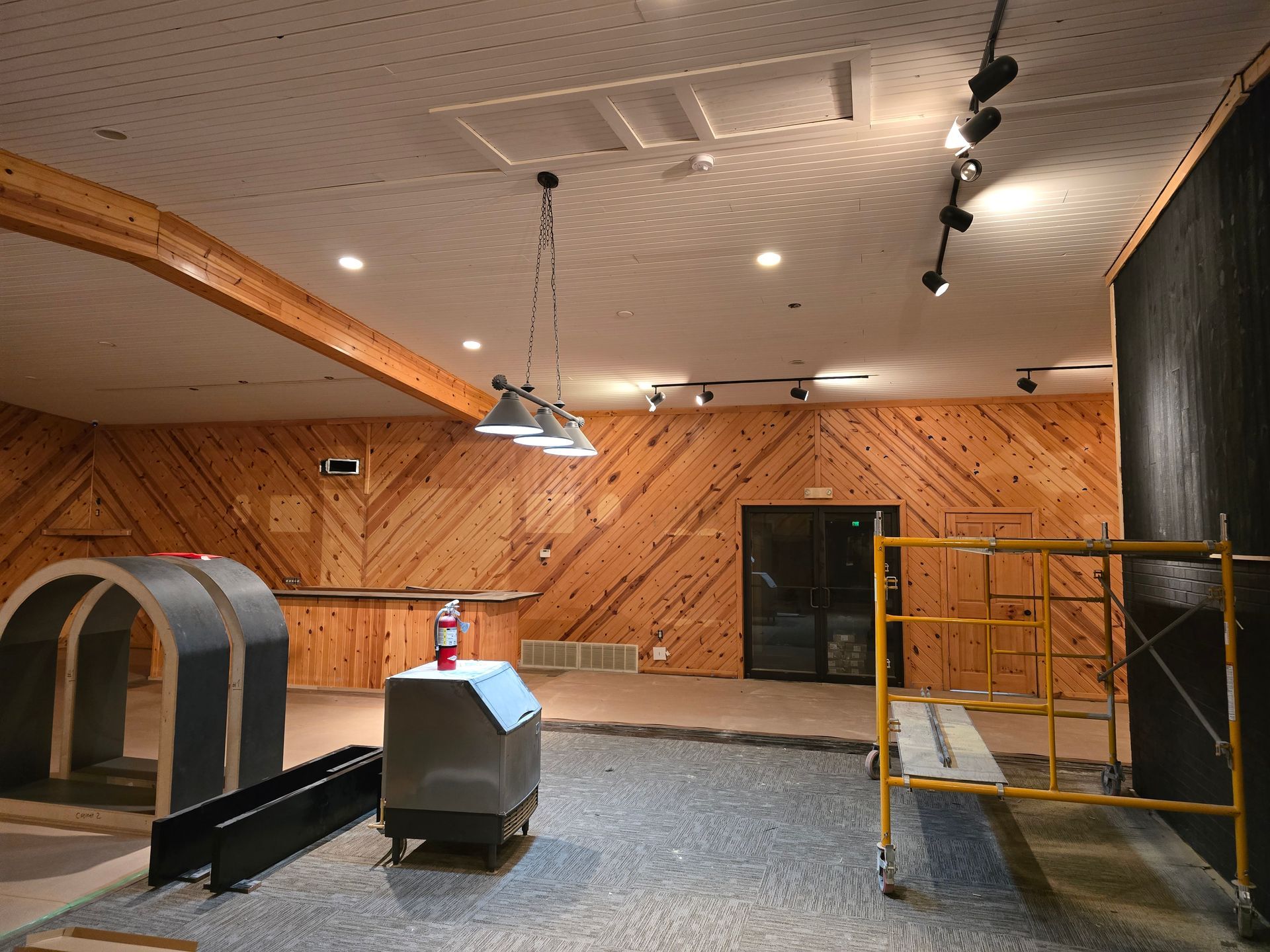 Interior of a room under renovation. Wooden walls and ceiling, construction debris on floor, scaffolding, and lighting fixtures.