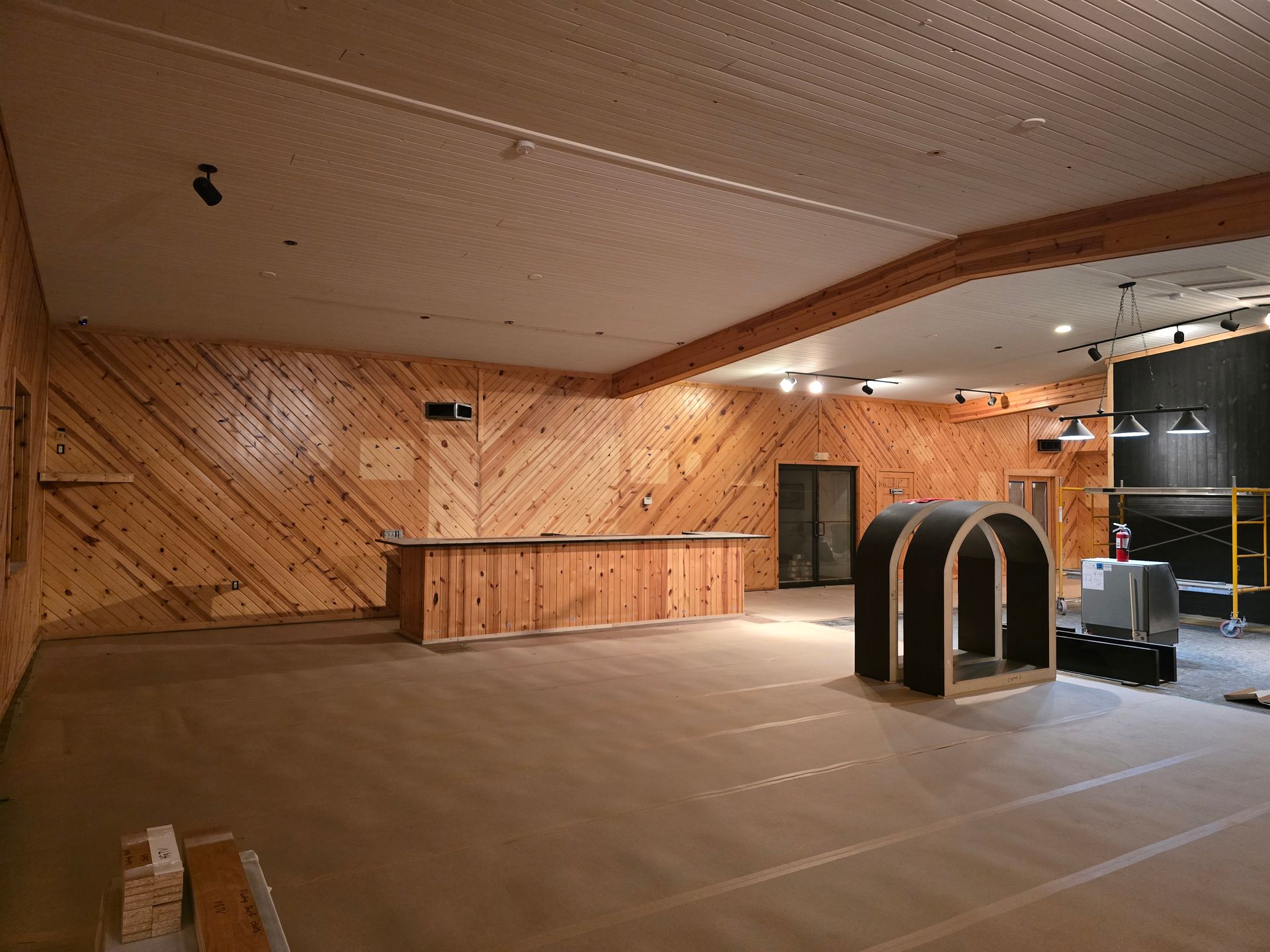 Interior of a room under construction with wood paneling and a bar, a dark arched structure, and exposed ceiling.