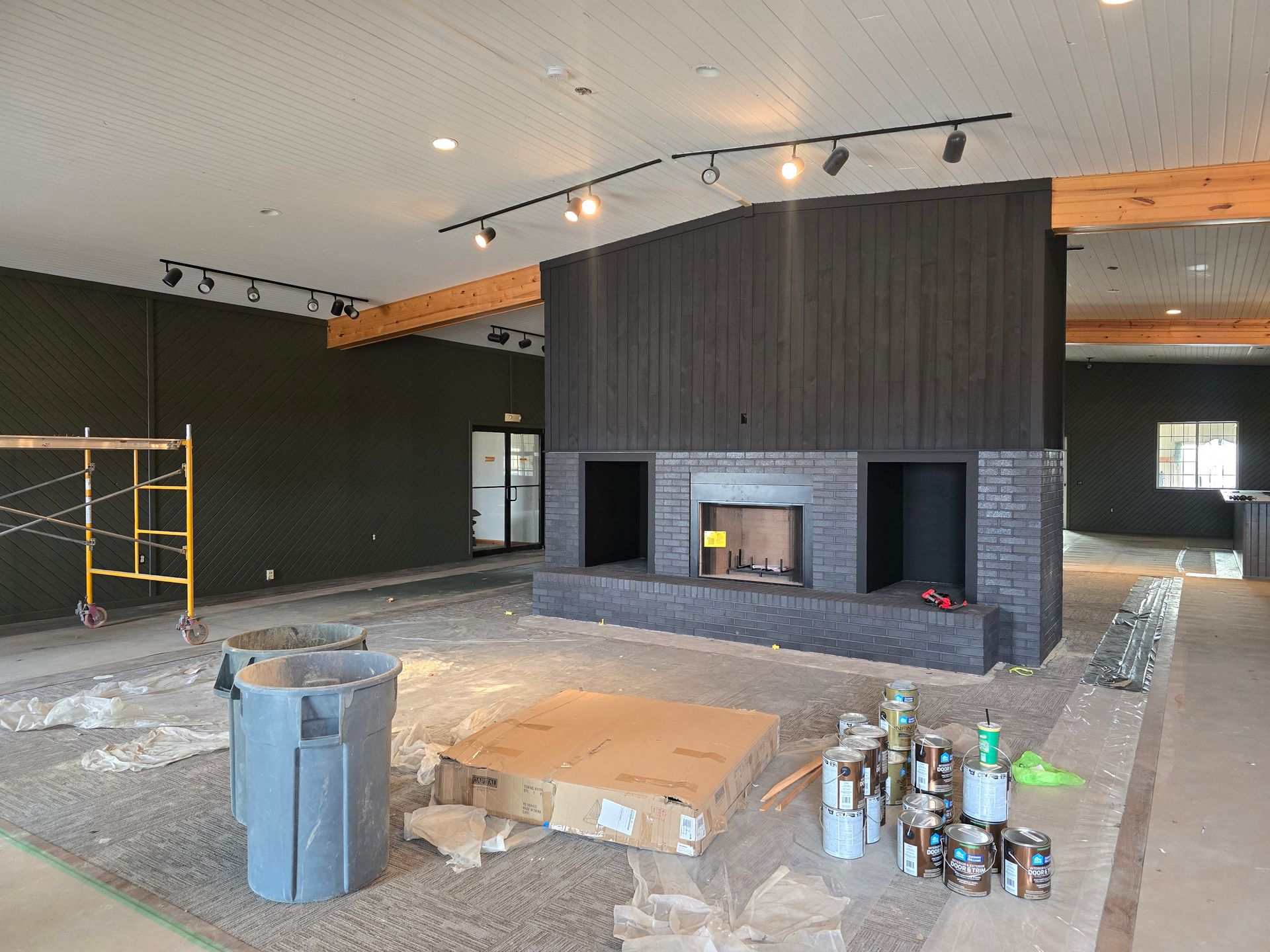 Interior under construction with dark fireplace, walls, and exposed beams. Scaffolding, debris, and cans of paint present.