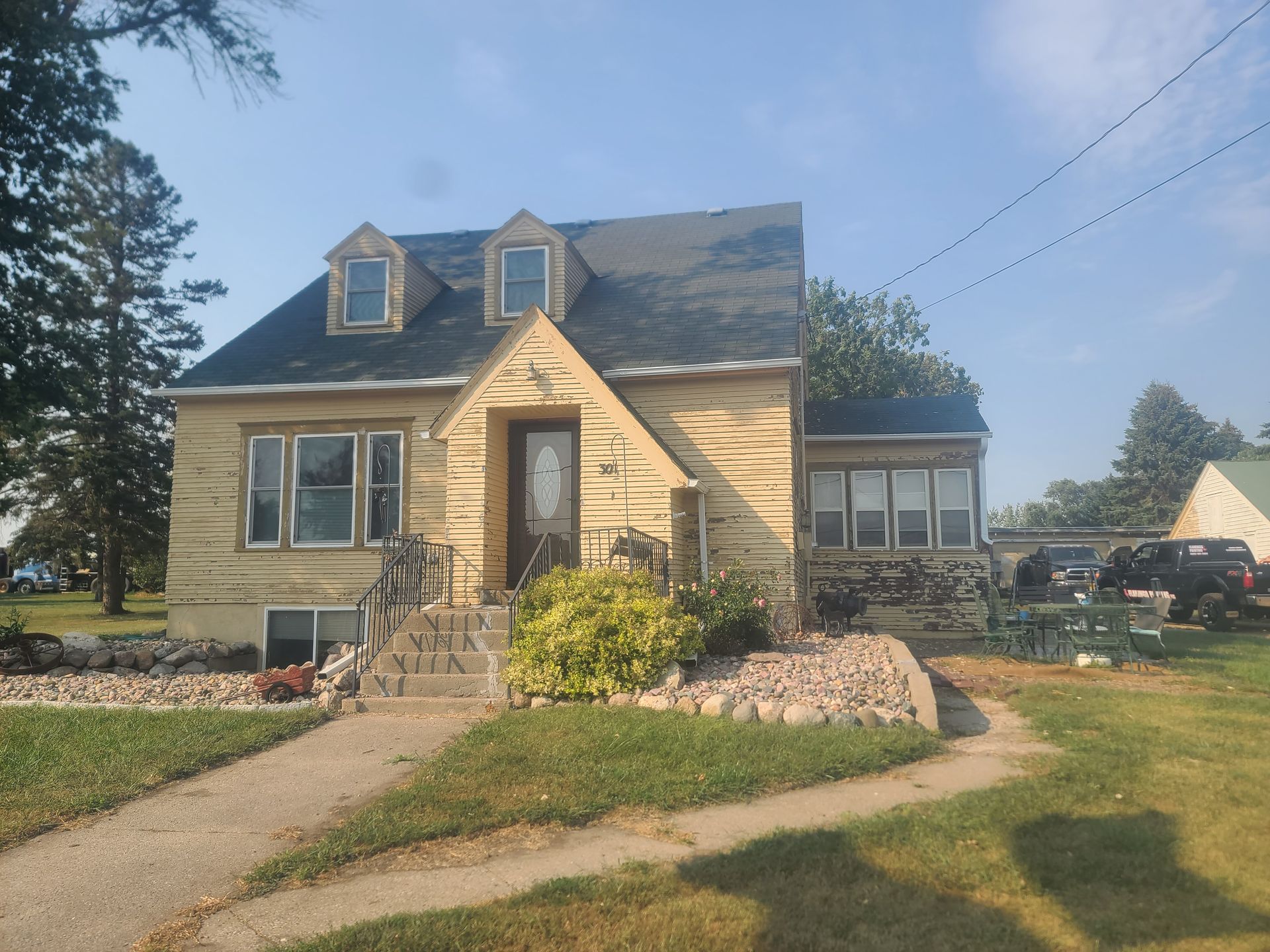 Yellow house with gray roof, three dormers, stone steps, and a concrete driveway.