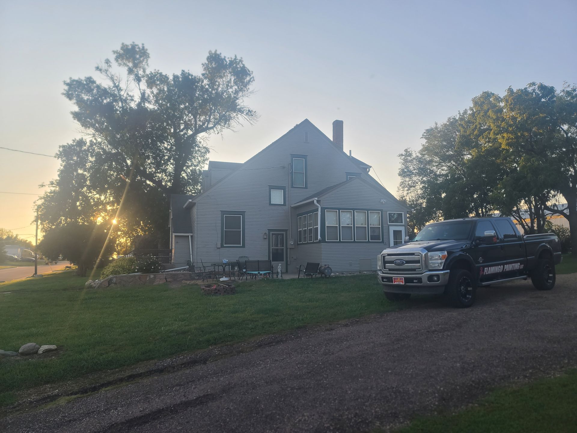 House with siding and a dark pickup truck parked in front; trees and the sunset are visible.