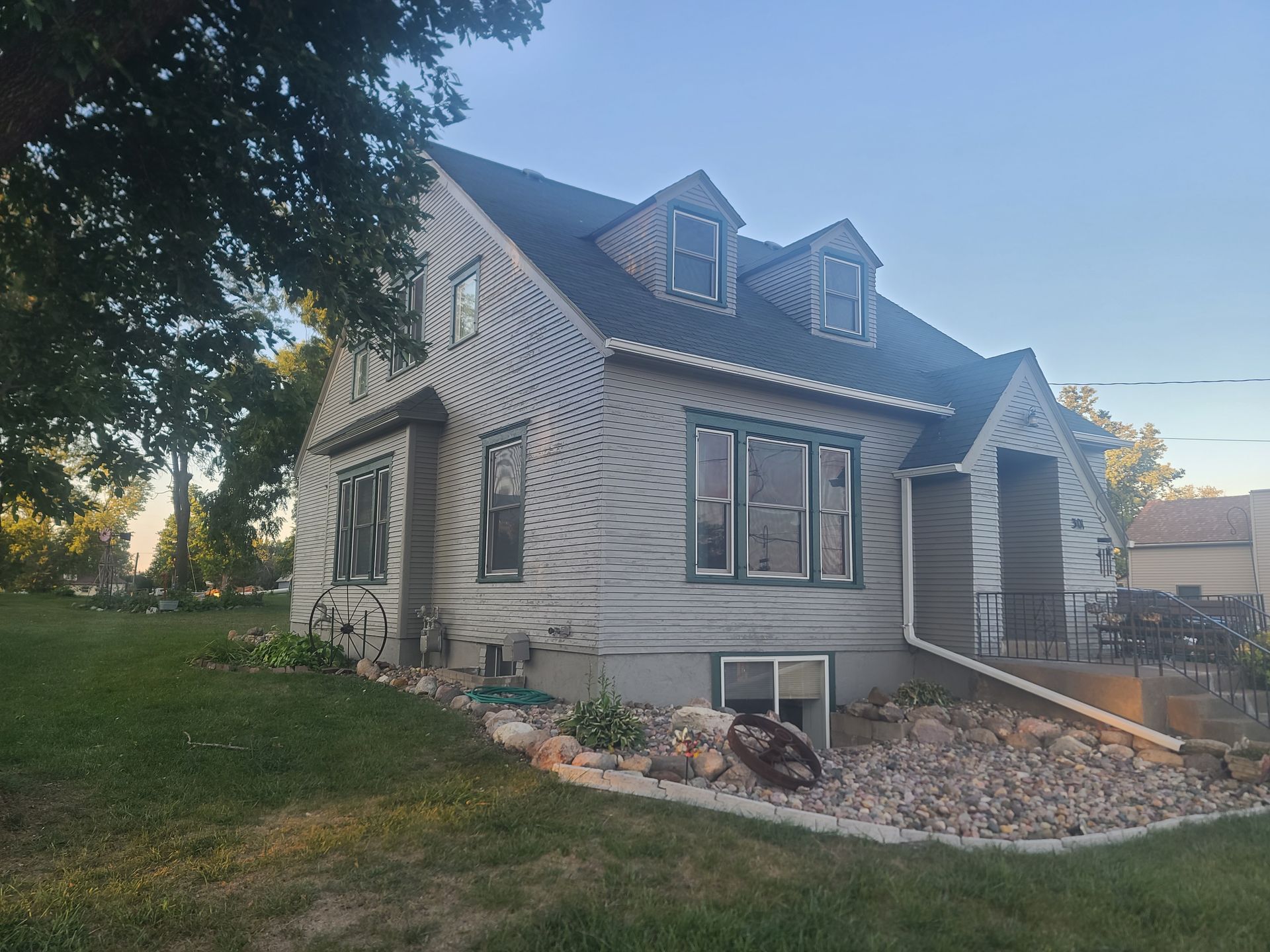 Gray house with dark roof and green window trim; front yard with grass and rocks.