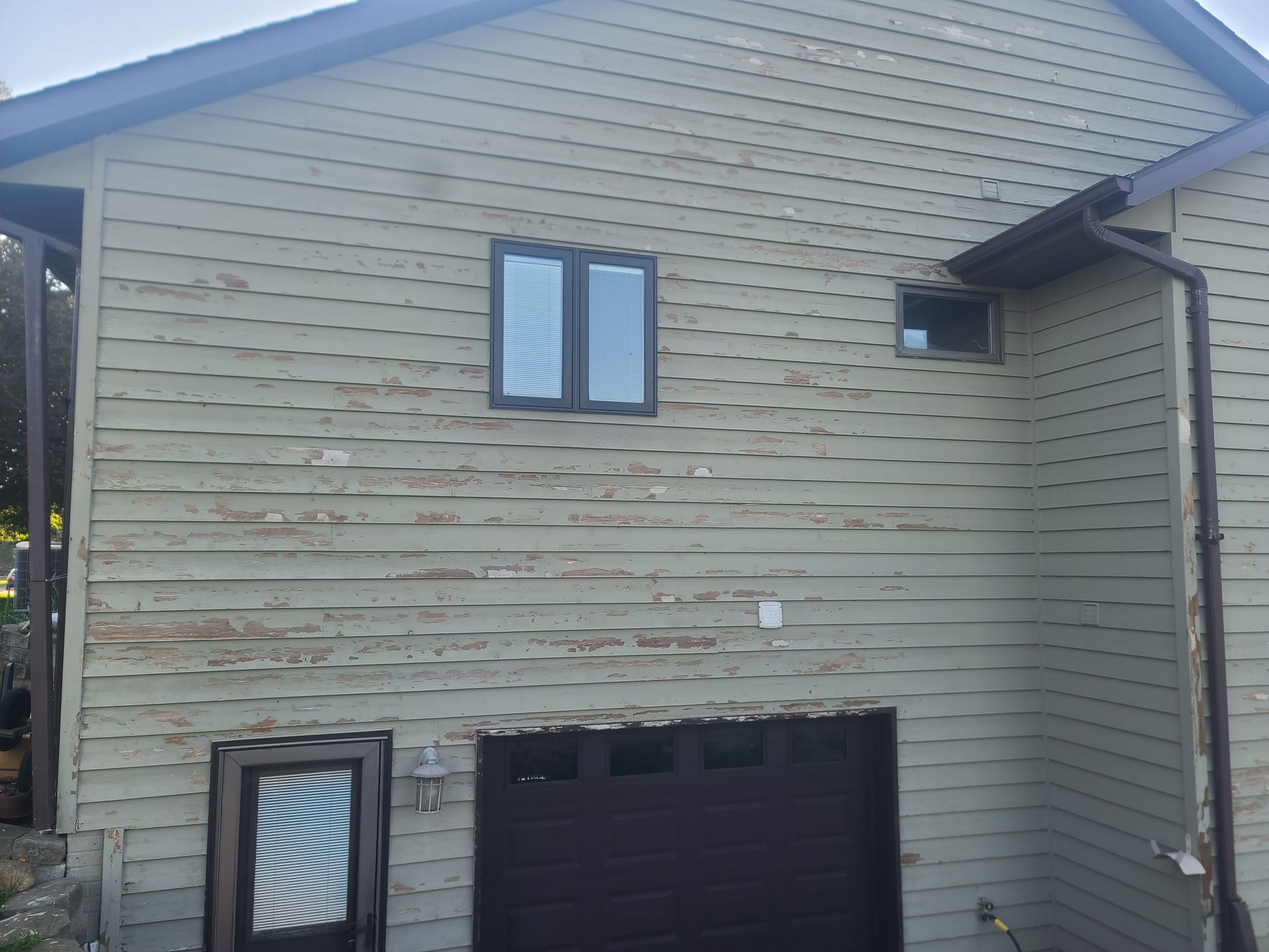 Exterior of a two-story building with peeling green siding, a garage door, and two windows.