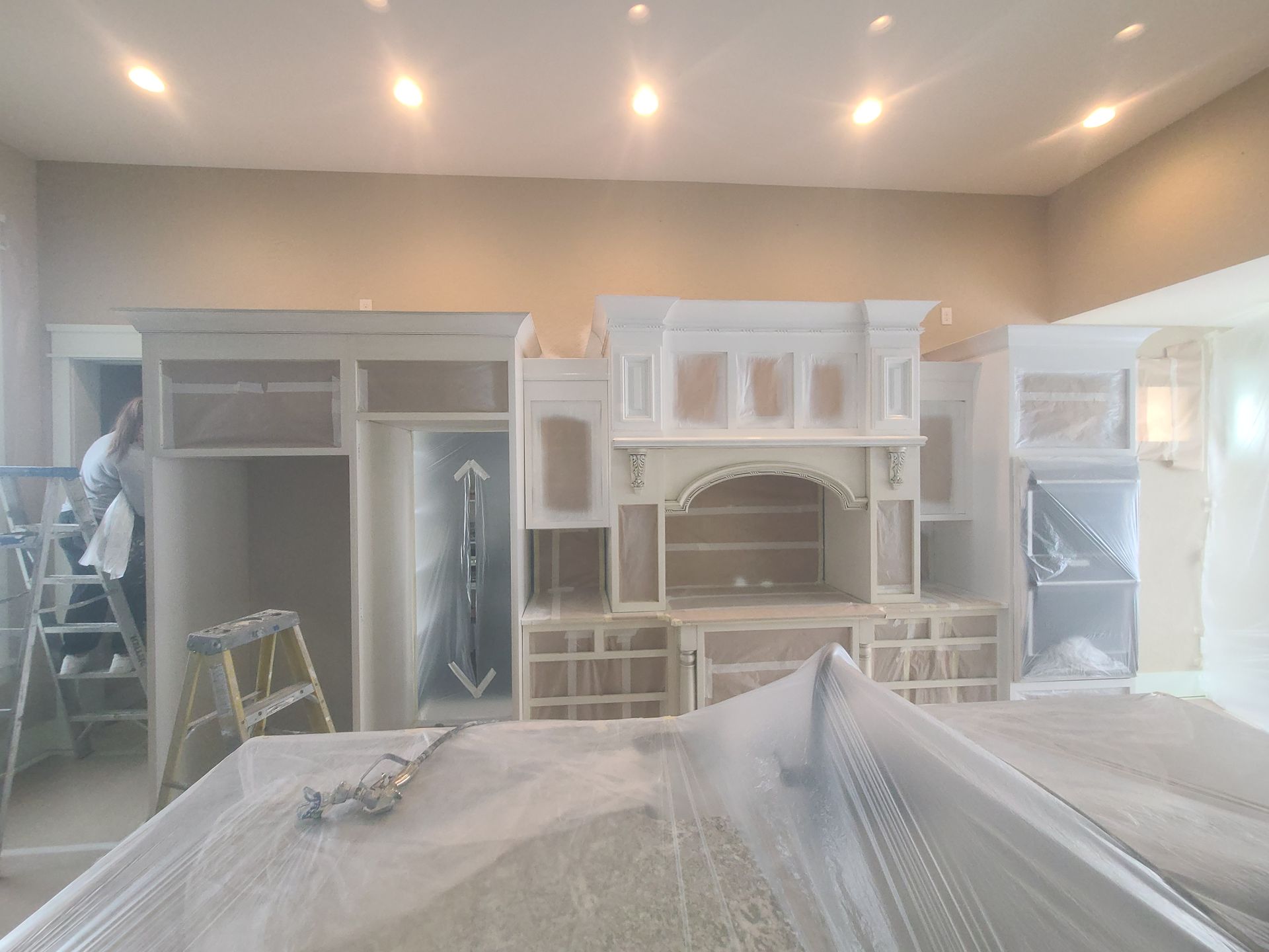 Kitchen cabinets being painted white; covered surfaces; worker on a ladder.