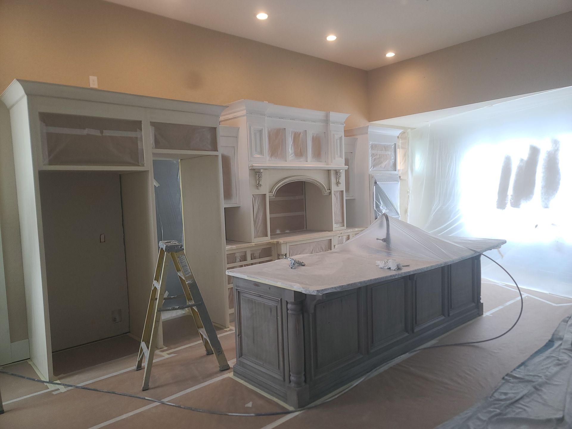 Kitchen cabinets and island being painted; plastic sheeting protects surrounding area.