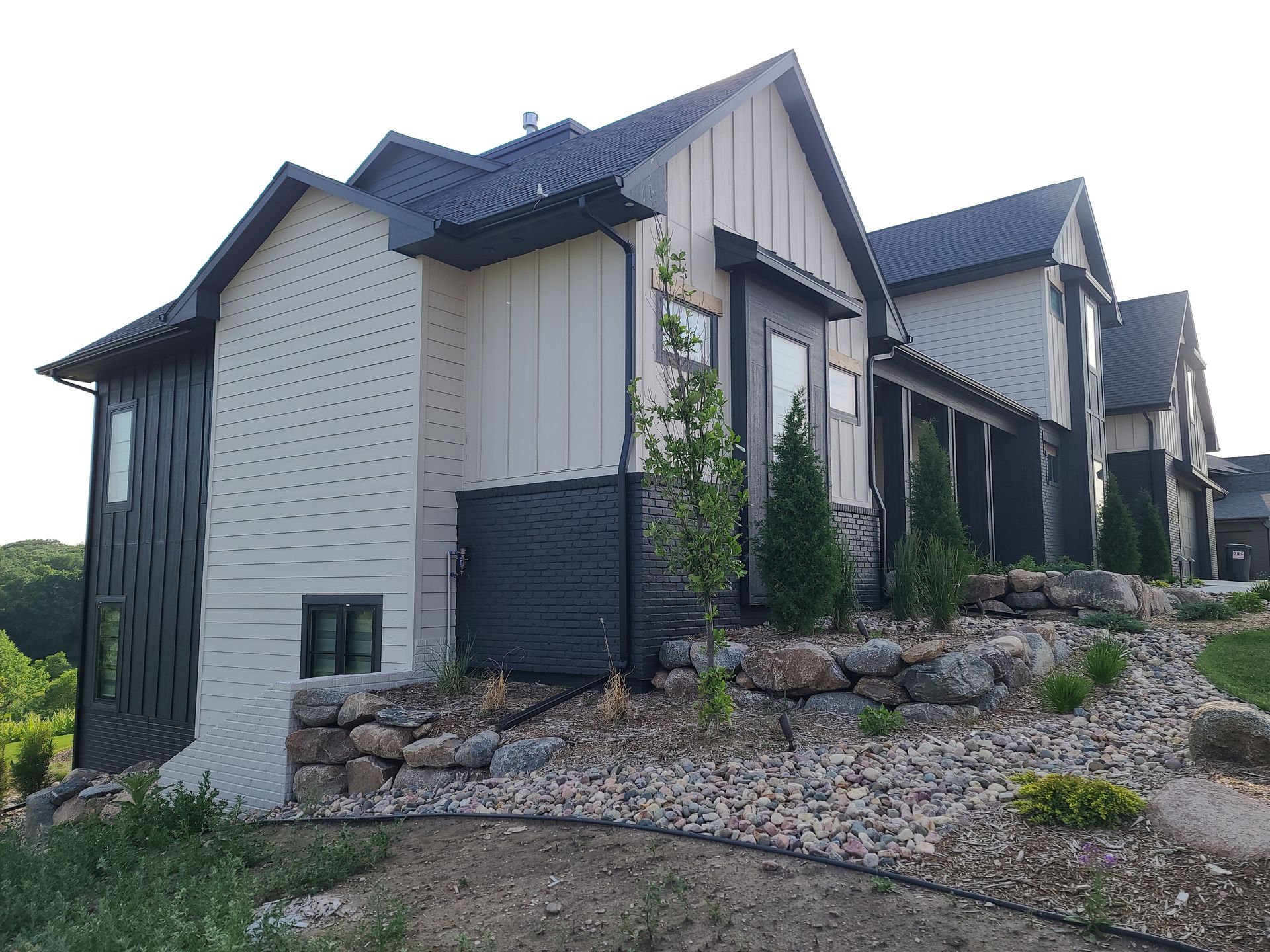 Modern house with black and tan siding, on a hill with landscaping and rock retaining wall.