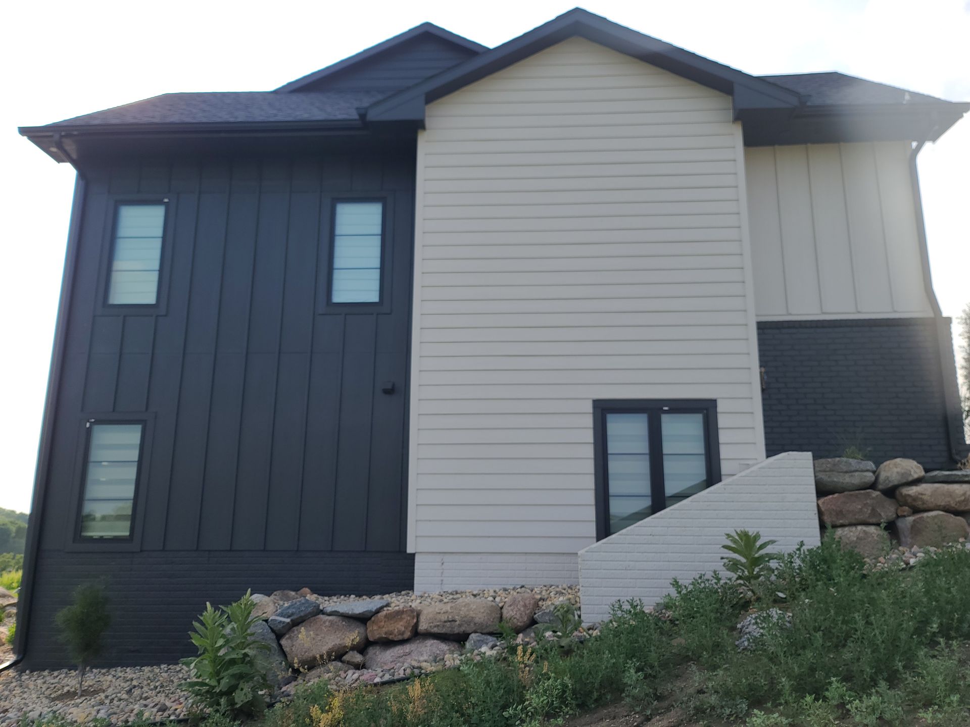 Two-story house with dark gray and beige siding. Small windows and a stone retaining wall.