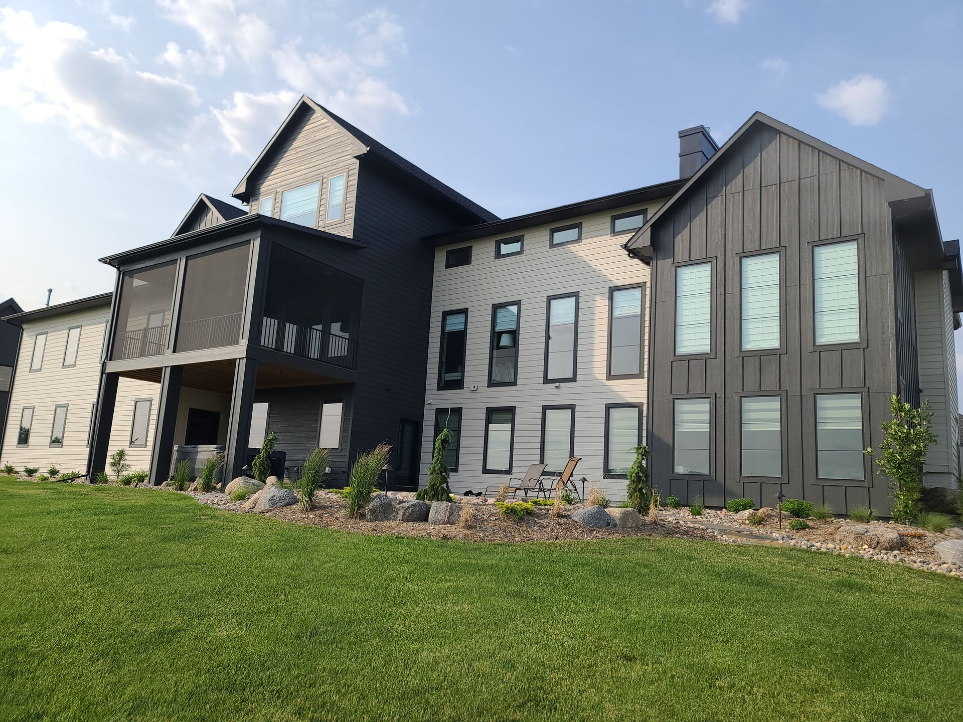 Modern two-story house with dark gray siding, light accents, and tall windows on a grassy lawn under a blue sky.