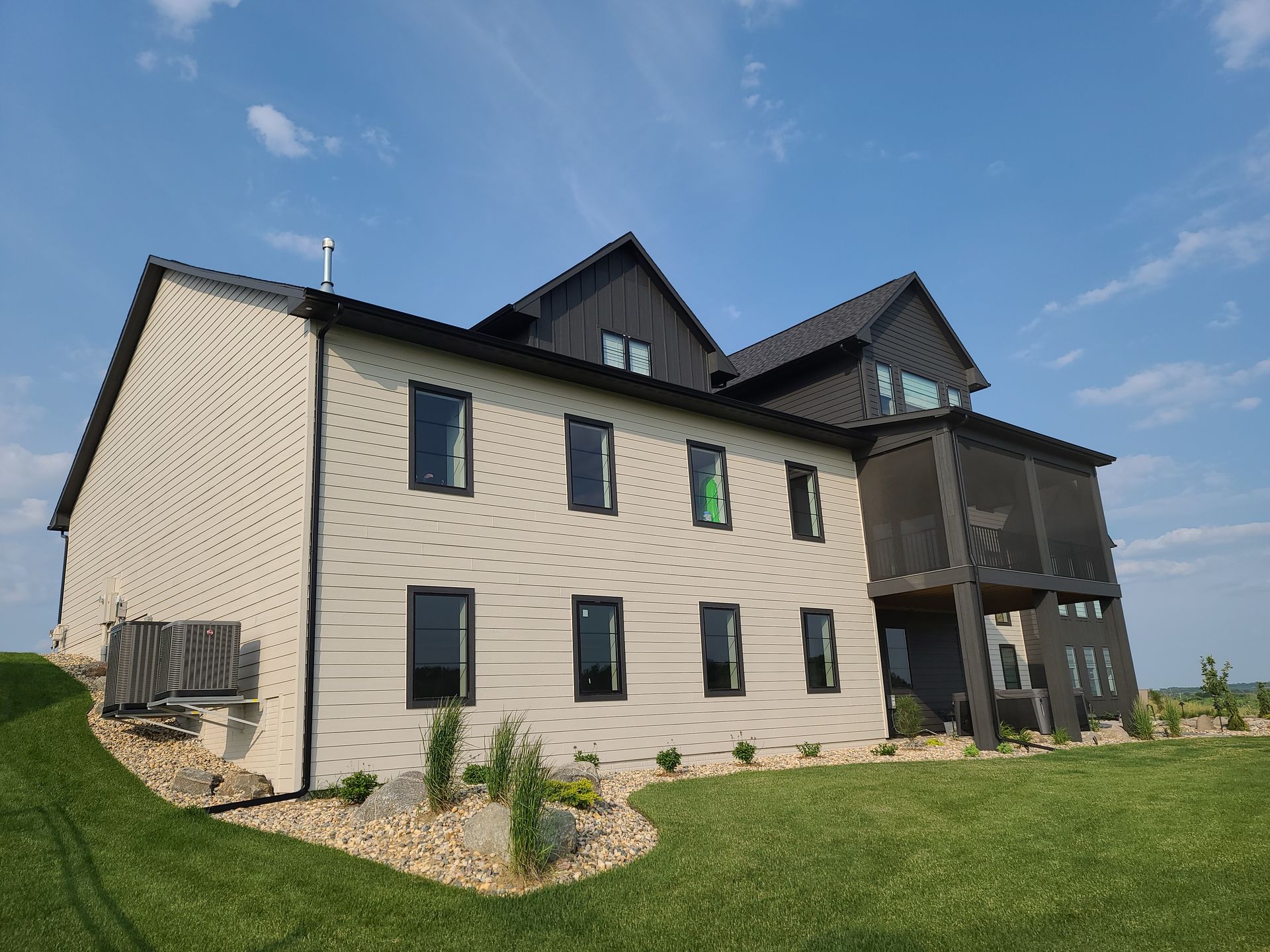 Two-story modern home with light siding, dark roof, and screened porch, set on a green lawn against a blue sky.