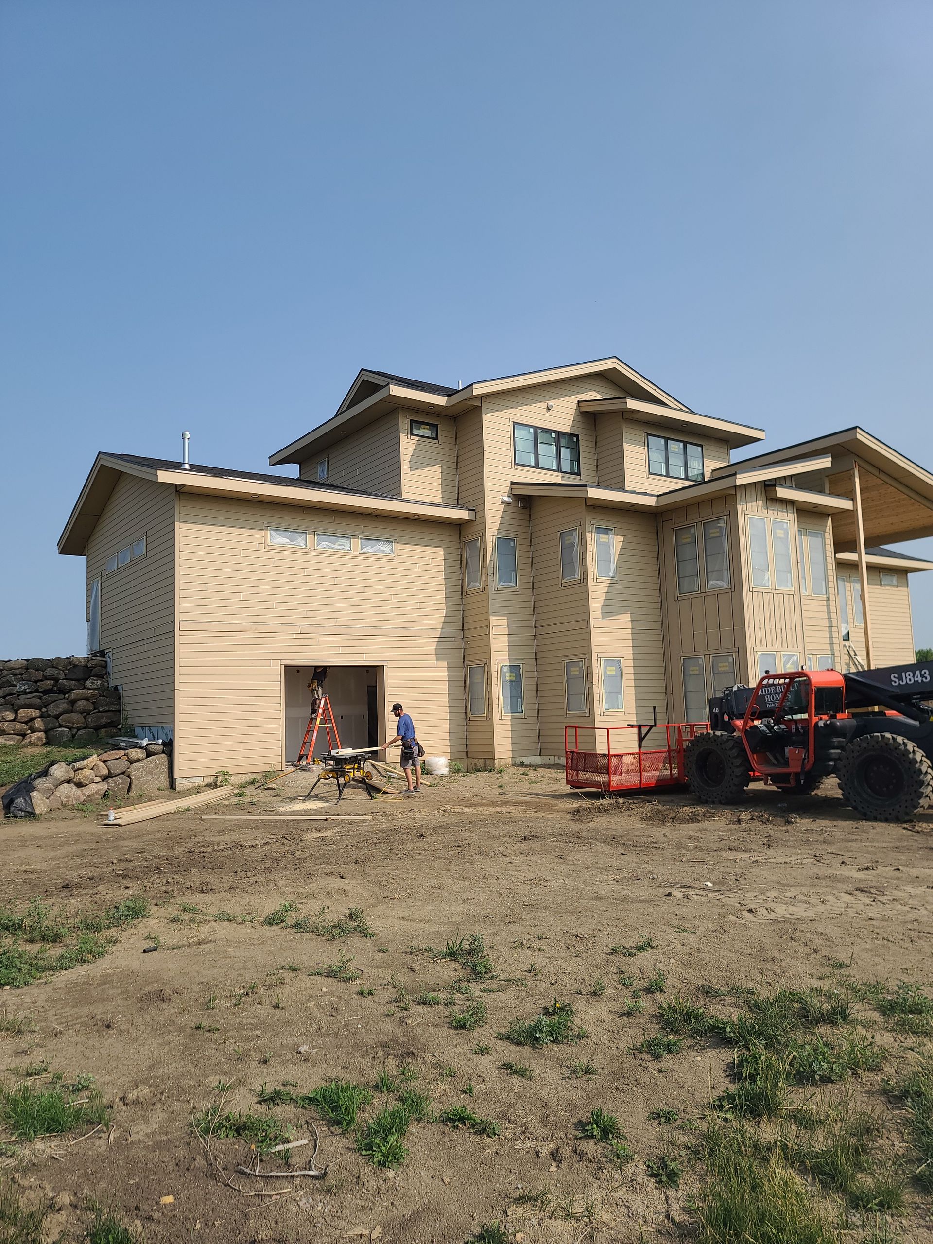 House under construction with workers, lift, and tractor on a hillside under a blue sky.