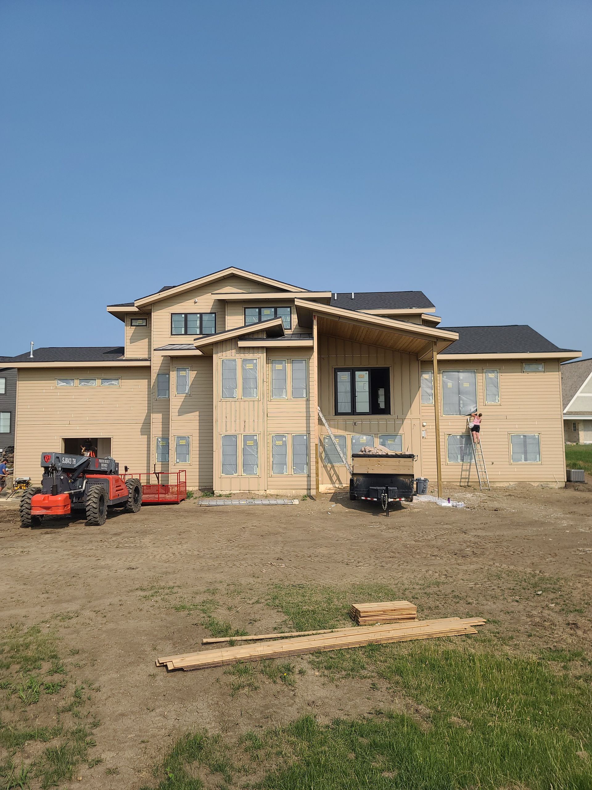 New house under construction, with wooden siding, windows, and roof. A tractor and trailer are present.