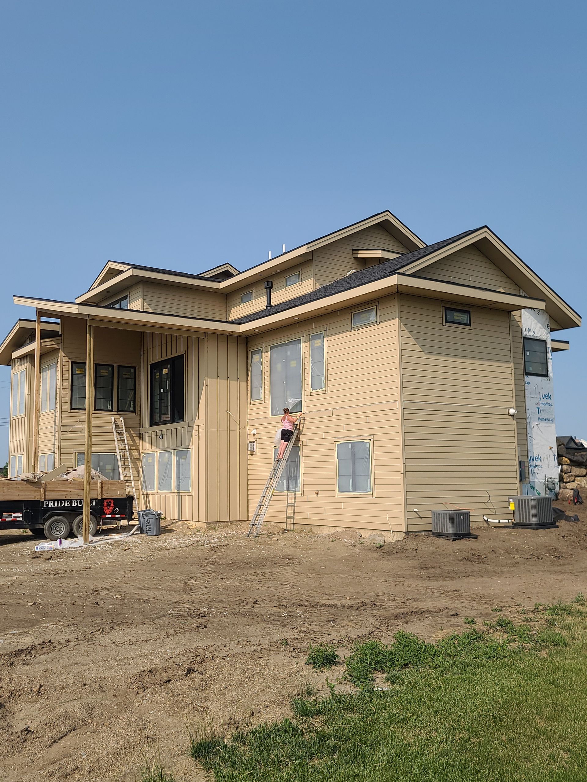 House under construction; light tan siding, worker on ladder painting, blue sky background.