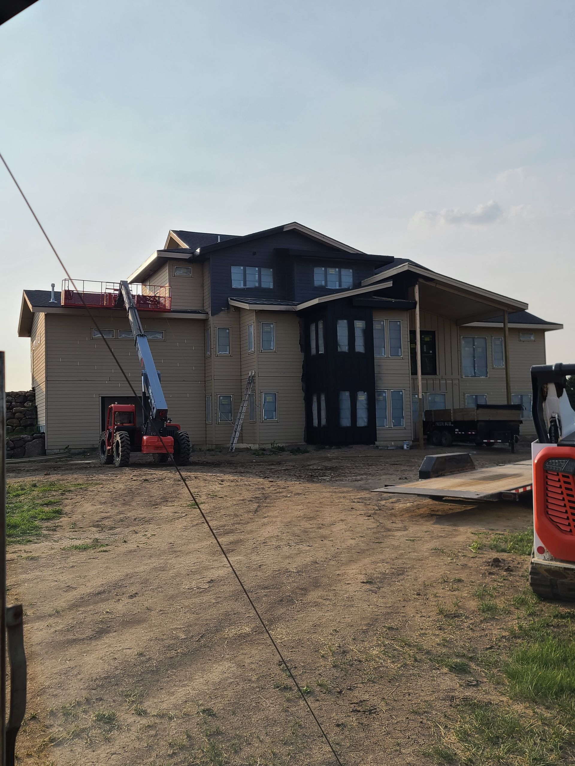 House under construction with an elevated lift; exterior beige siding, dark trim, and a dirt yard.