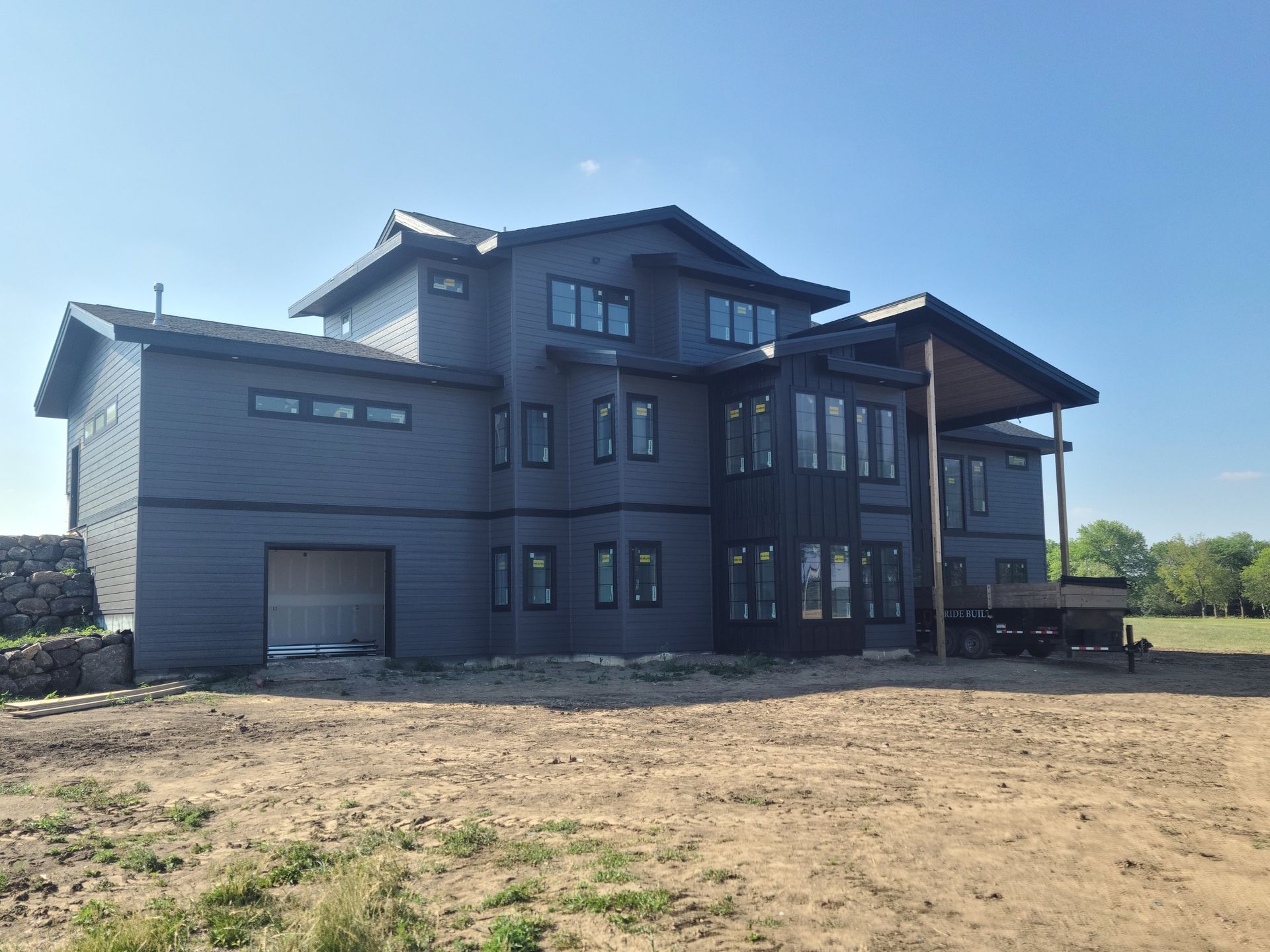 Dark blue modern house with multiple windows and a covered porch on a dirt lot.