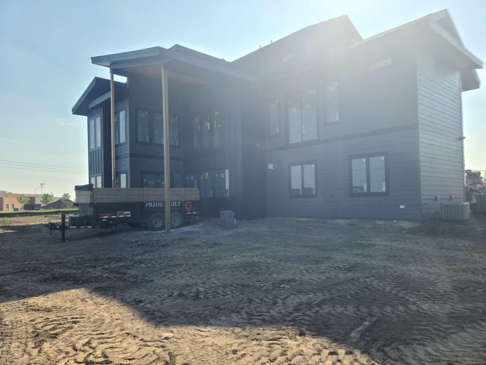 Dark-sided house under construction with a deck, windows, and a sunny sky background. Dirt ground in front.