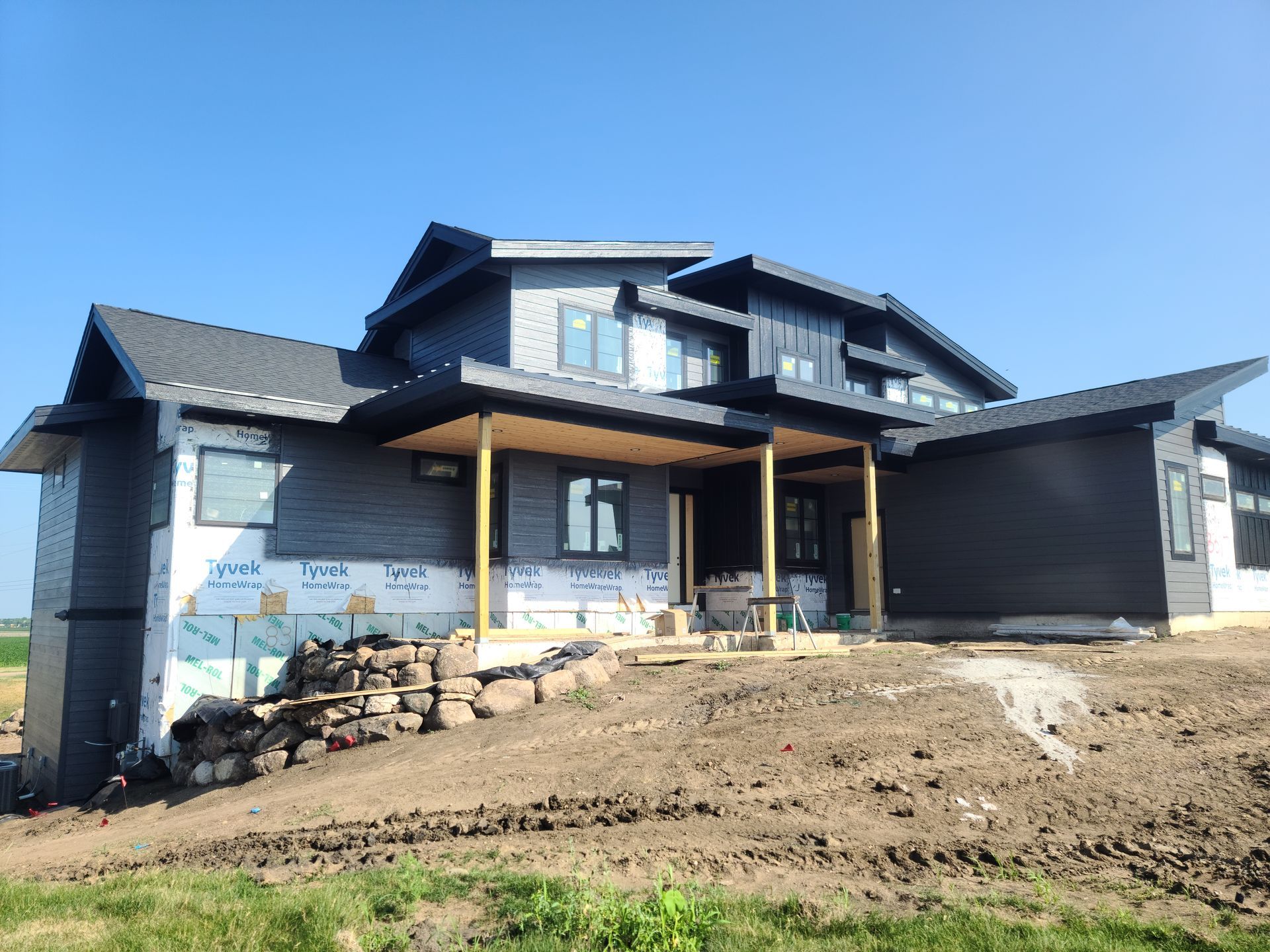 Modern house under construction; dark gray siding, black trim, blue wrap, on a sloped dirt lot, blue sky.