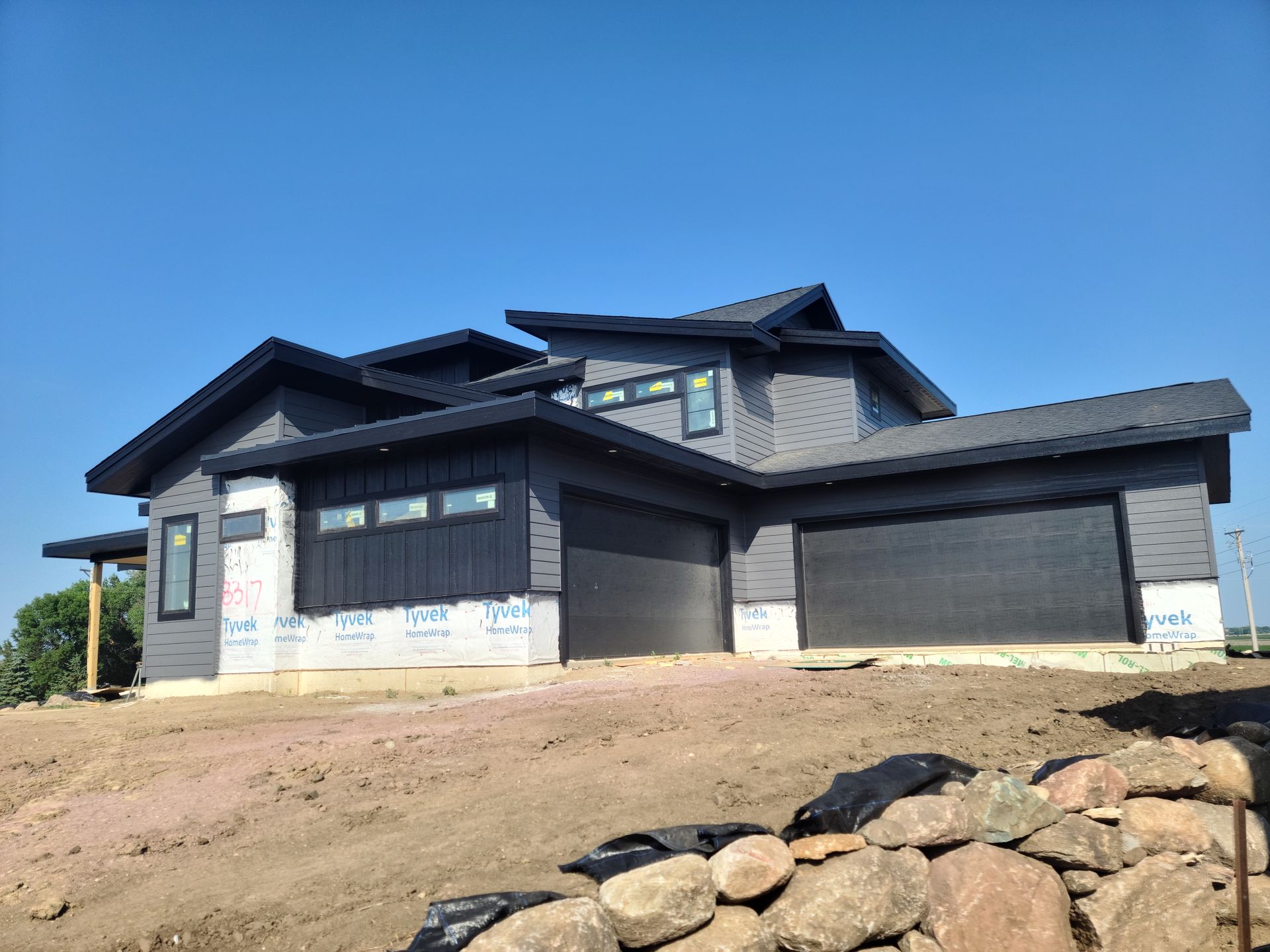 Two-story modern house under construction; gray siding, black trim and garage doors, blue sky, and dirt.