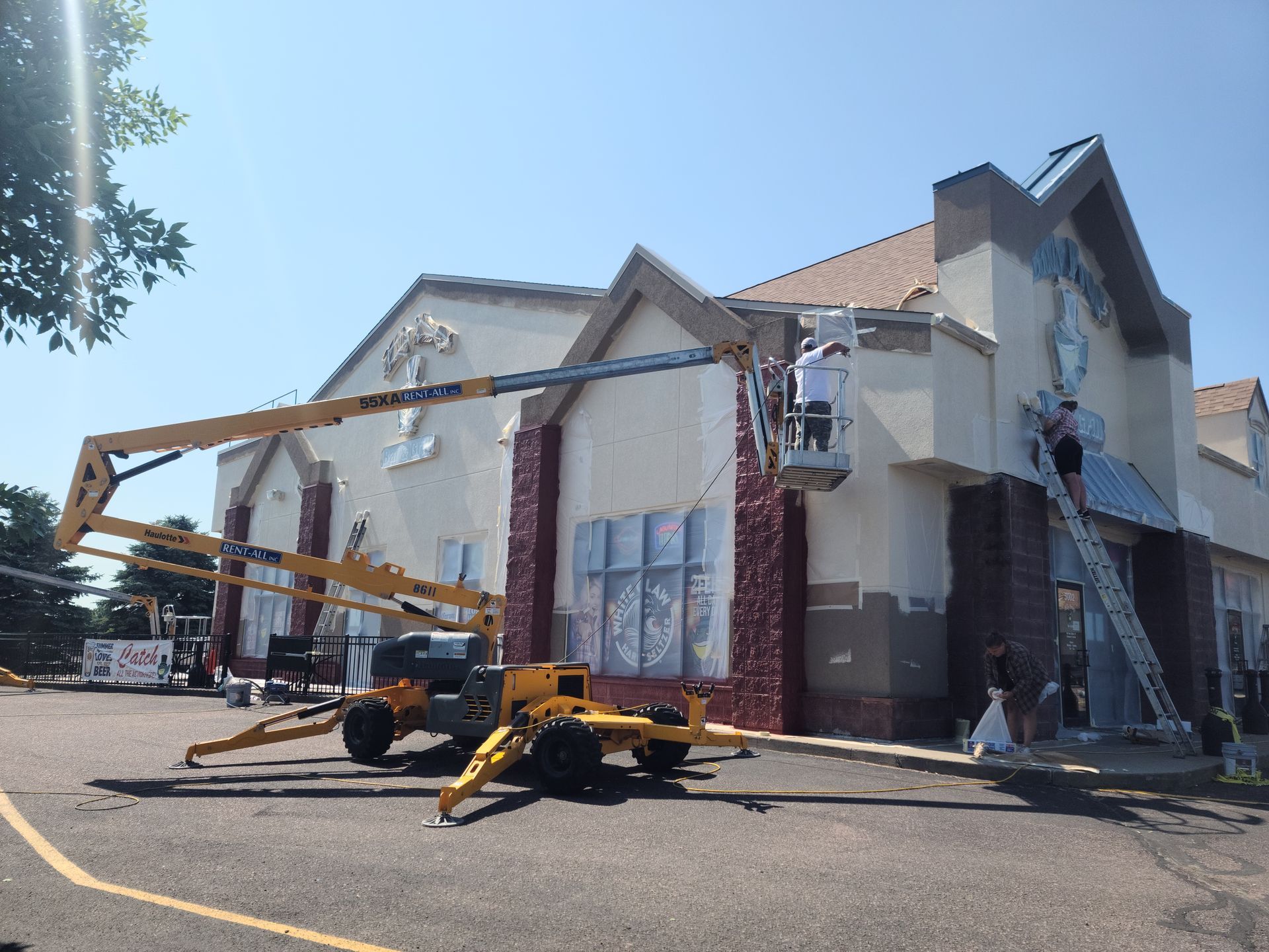 Building exterior being painted, with two workers in lifts and one on a ladder. Yellow and brown machinery.