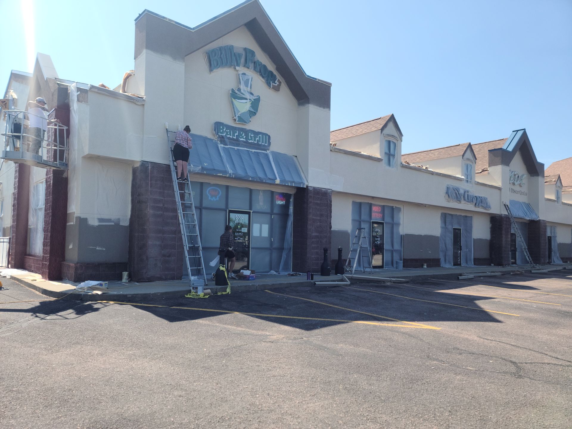 Exterior of a building under renovation with workers on a ladder, storefronts, and a parking lot.