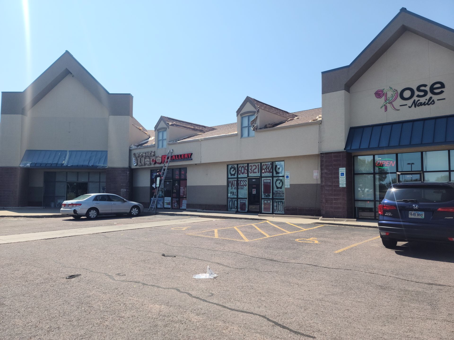 Exterior of a shopping center with Rose Nails and other stores on a sunny day. A car is parked out front.