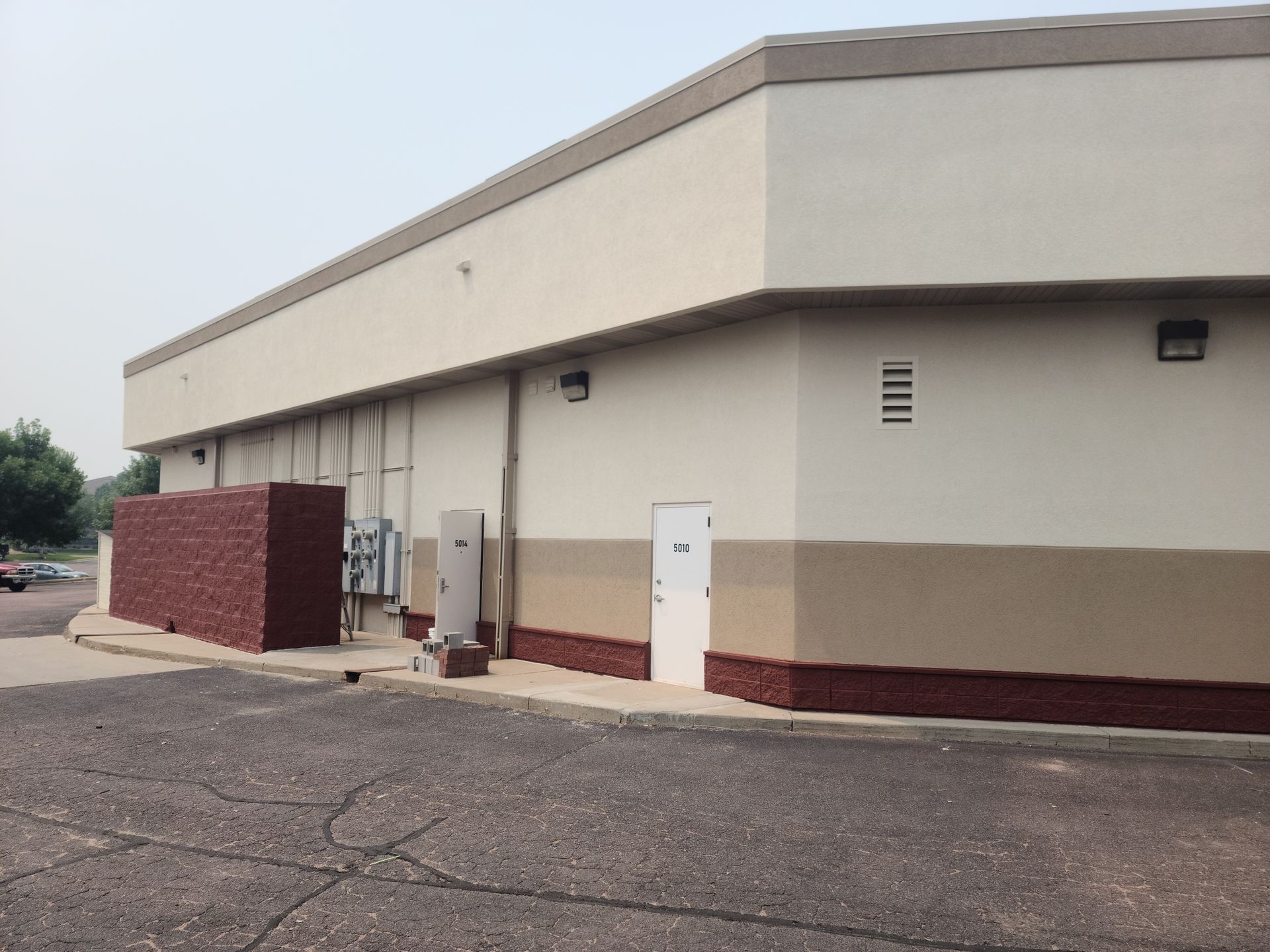 Exterior view of a commercial building with brick base, tan walls, and a light-colored roof. A doorway and utility boxes are present.