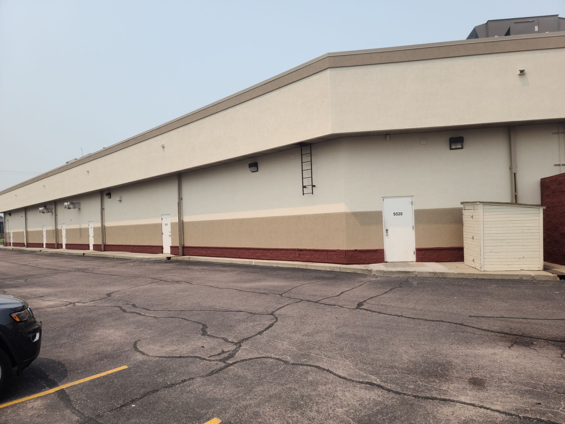 Exterior view of a long, light tan and brown commercial building with a cracked parking lot.