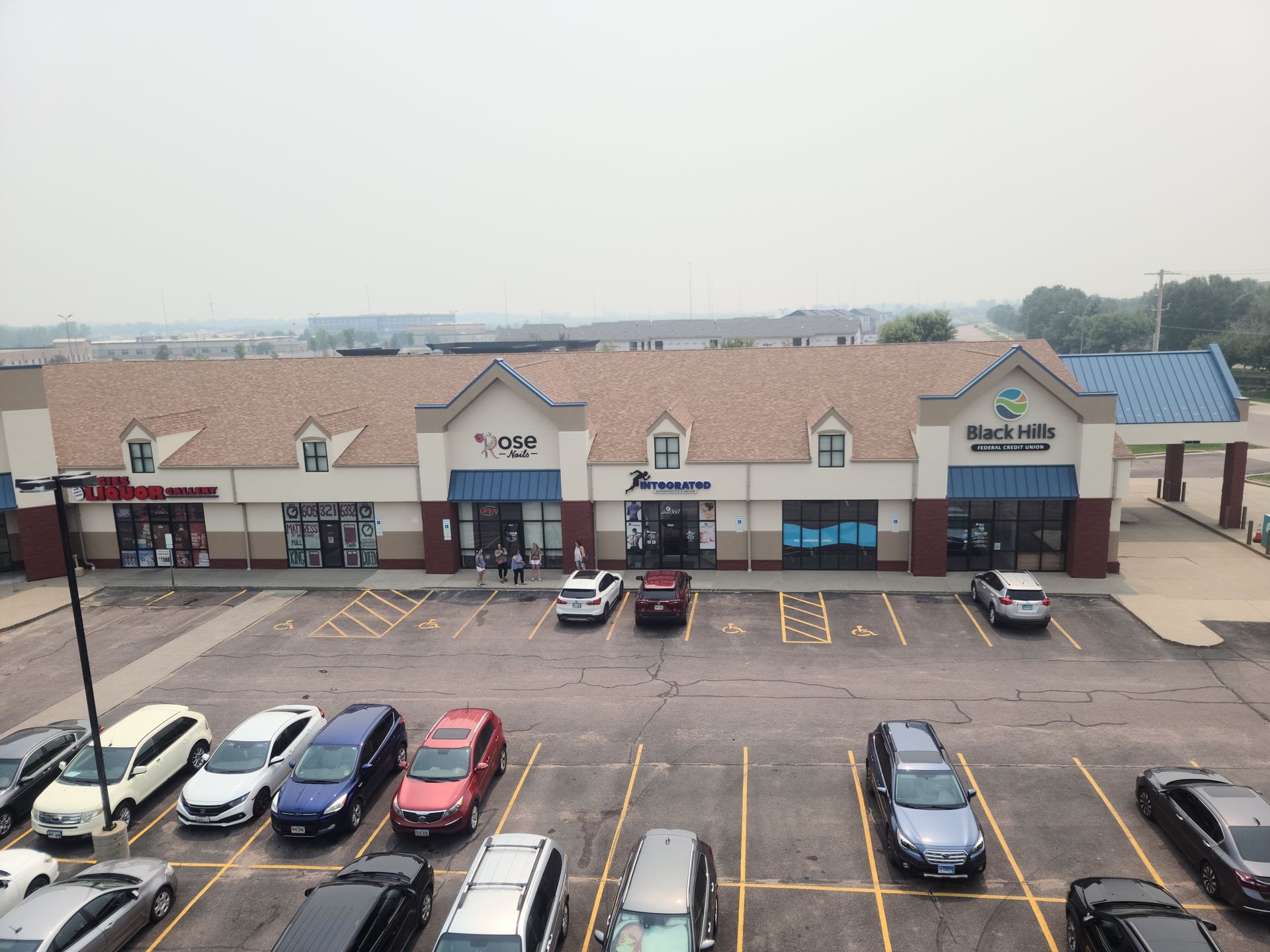 A strip mall with various storefronts, cars parked in front. Gray sky.