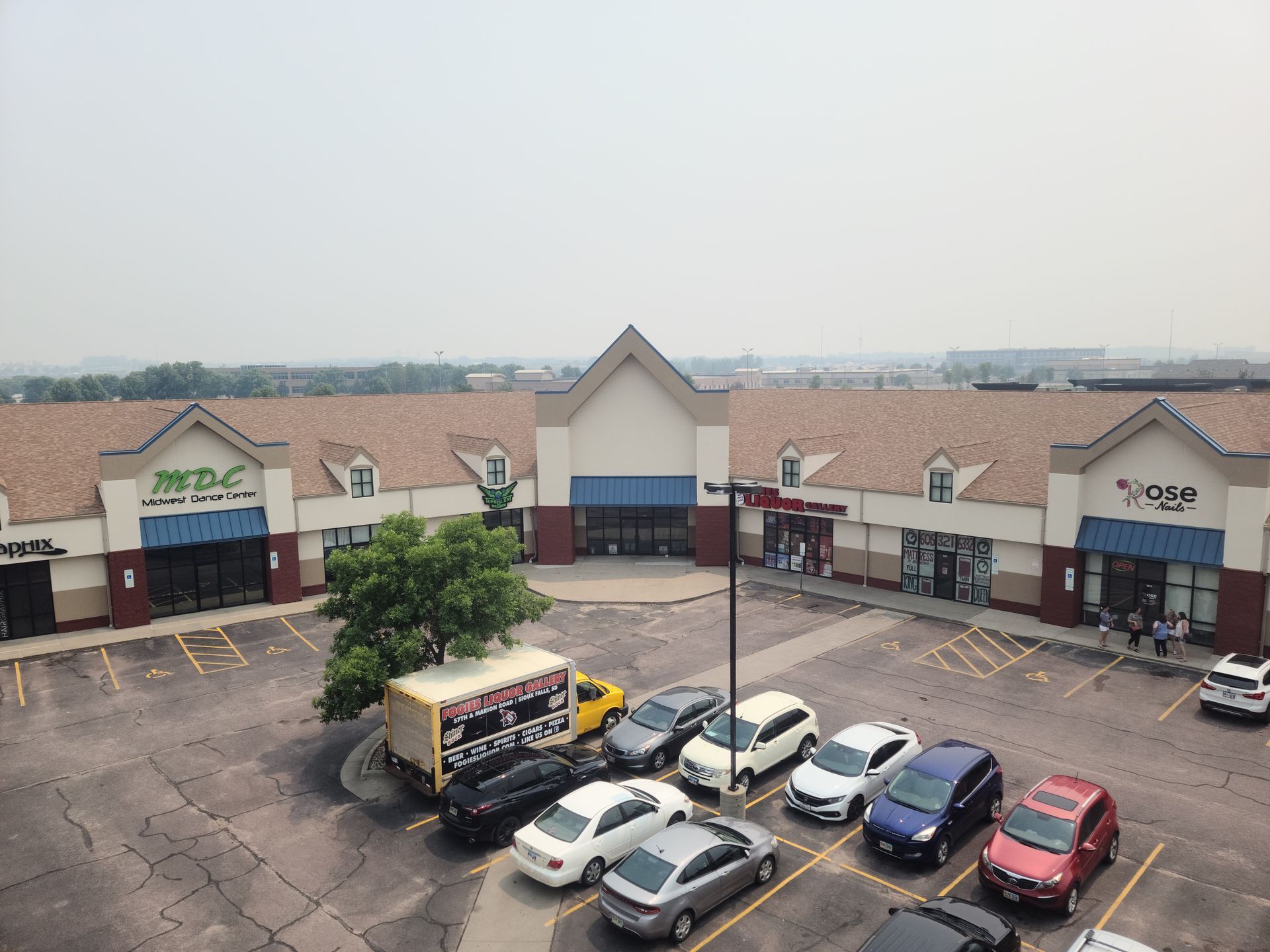 Shopping center exterior with parked cars. Buildings have brown roofs and tan facades. Overcast sky.