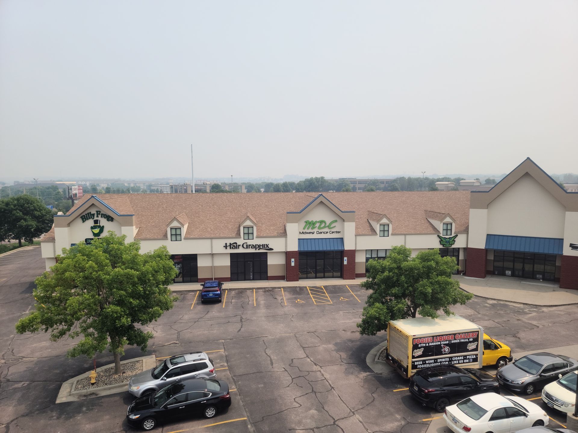 Exterior view of a shopping center with various stores, a parking lot with cars, and a hazy sky.