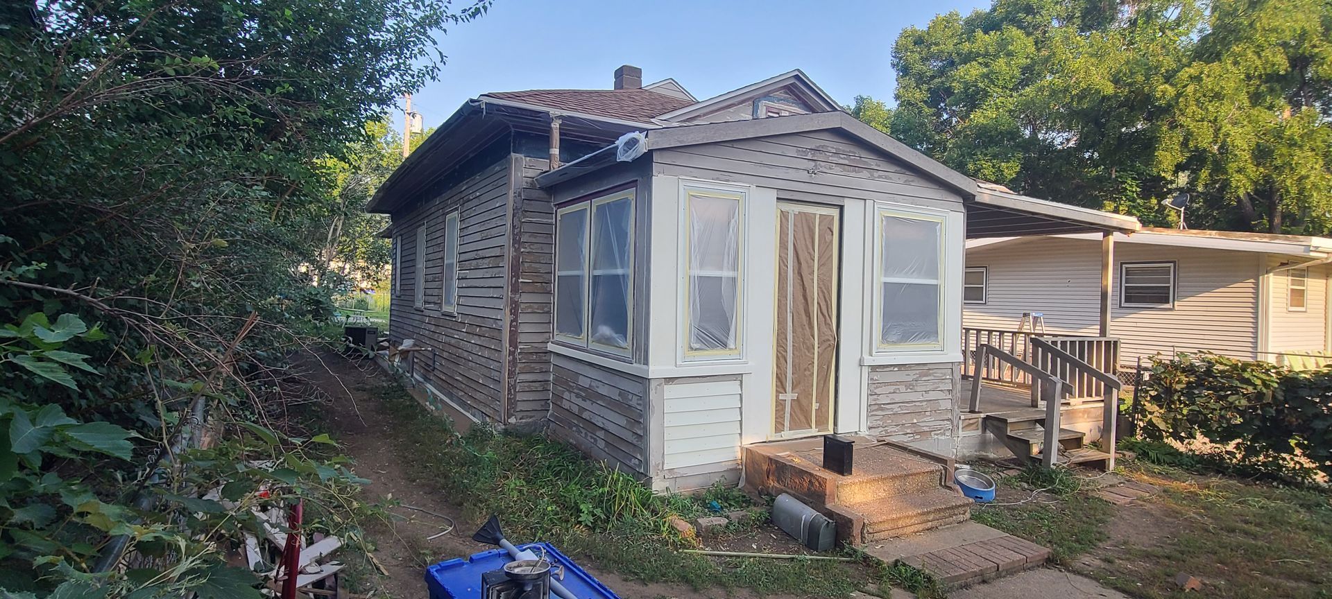 Dilapidated one-story house with overgrown yard, weathered siding, and boarded-up door.