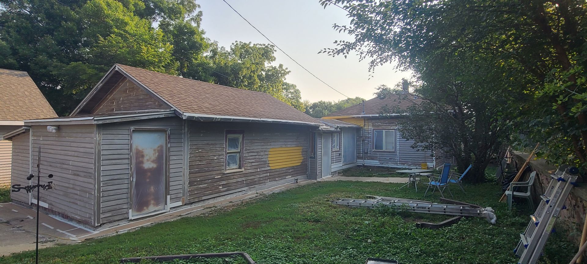 A weathered building with a brown roof and gray walls, in a yard with green grass and trees.