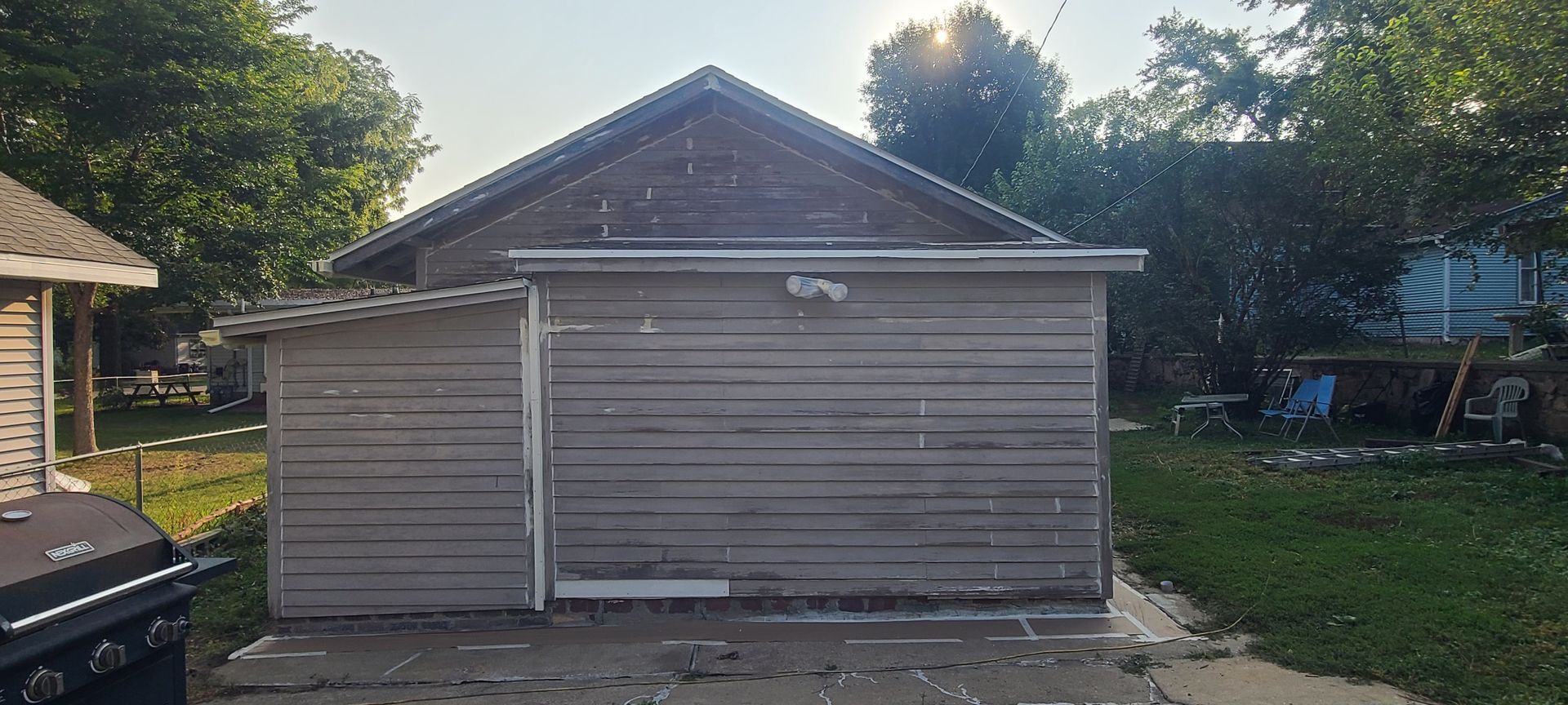 Gray garage and shed with trees in the background, sunlight peeking through.