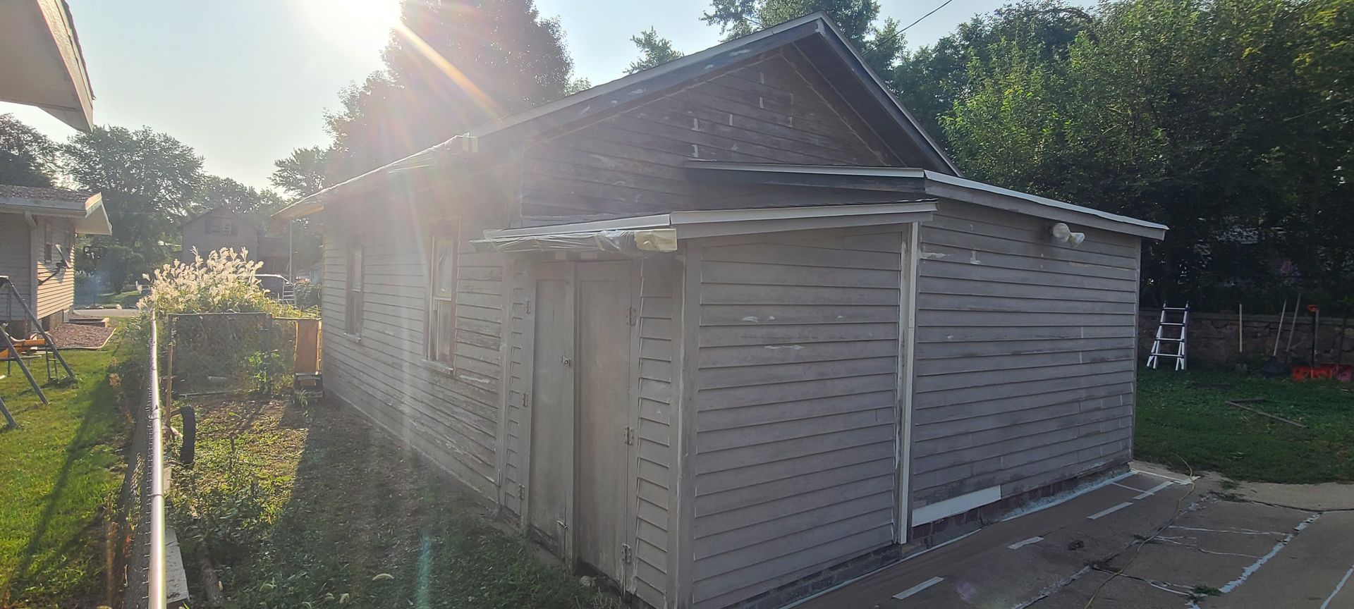 A weathered wooden shed with a dark roof stands on a grassy lot, sunlight streaming from above.