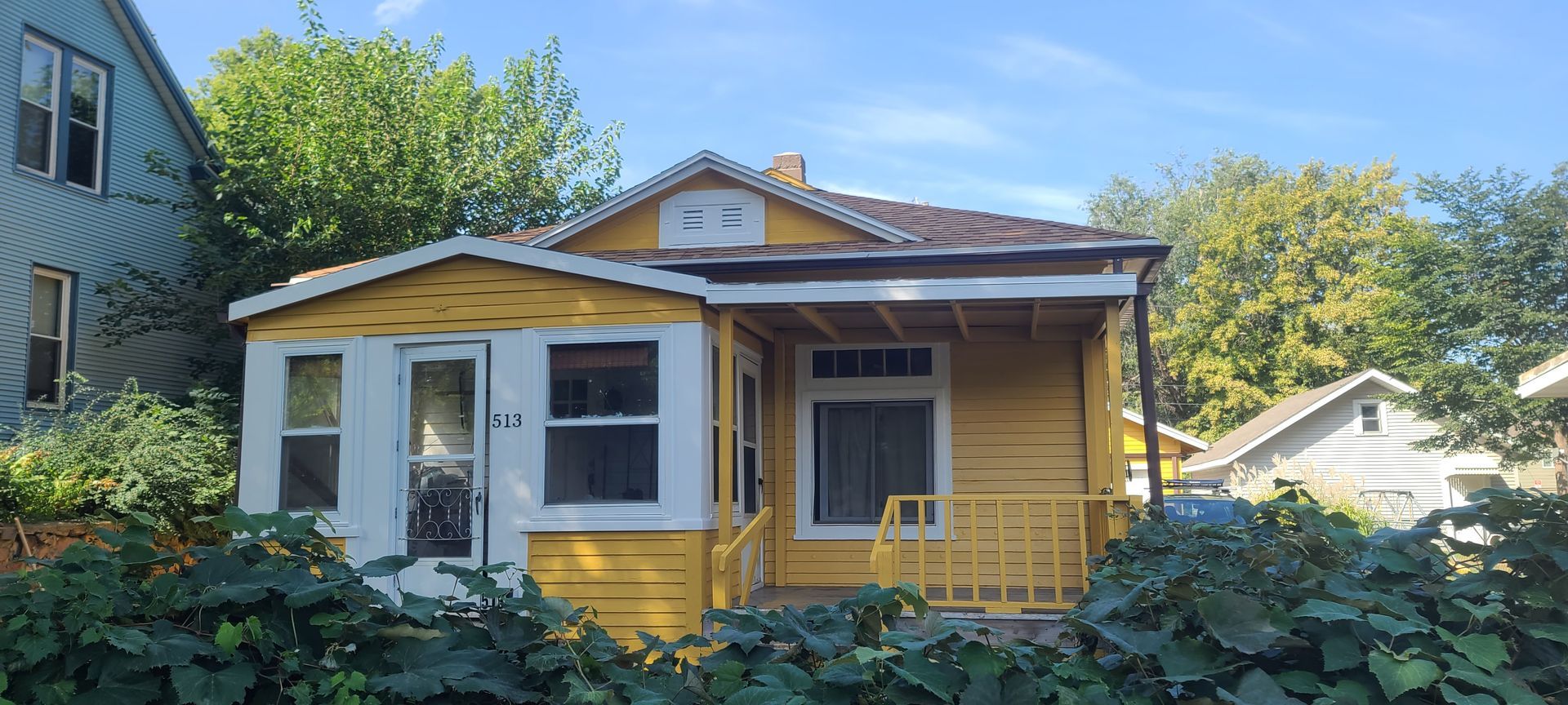 Yellow house with white trim and a small porch. Green foliage in foreground. Blue sky.